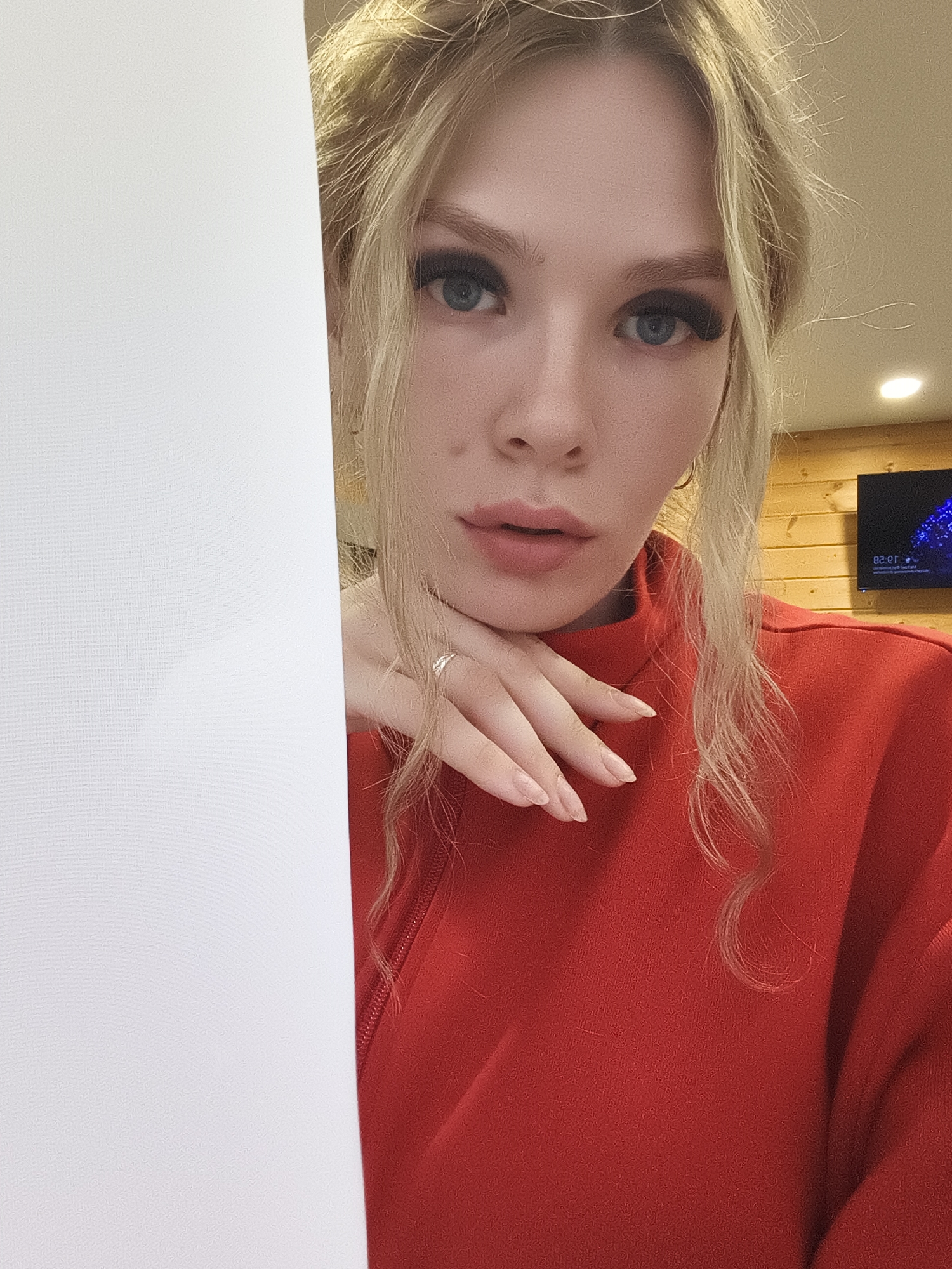 A close-up shot of a fair-skinned woman with blonde hair, wearing a red top, holding her chin with her hand. She has blue eyes and pink lips. She is wearing a silver ring on her finger. To her left is a white textured surface, partially obscuring the view. In the background, blurred, is a wood-paneled wall with a television showing a digital display. The time on the display reads "10:28." The location is likely inside a building, possibly in Русский Вожой, Russia.