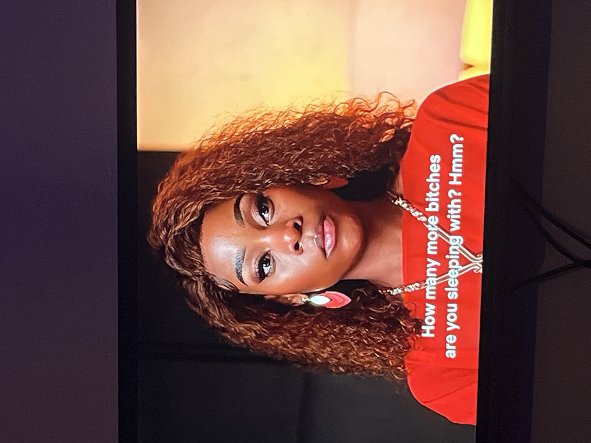 A young Black woman with curly, reddish-brown hair and light makeup is shown from the chest up, facing the camera with her head tilted to her left. She is wearing a bright red top and a delicate silver necklace. She has large, pink, leaf-shaped earrings. Her expression is neutral, with her eyes looking directly at the viewer.
Text is visible on her red top, written vertically in white letters: "How many more bitches are you sleeping with? Hmm?". The background is a soft, yellowish-orange gradient.
The image appears to be a screenshot from a video or television program, given the dark edges on the right and left sides that suggest a screen frame. The lighting is bright and focused on the woman's face, creating a warm glow. There are no obvious clues about the time of day or weather. The context of Gura Top, Nigeria, is provided but not visually evident in the image itself. The overall mood is somewhat provocative due to the text displayed.