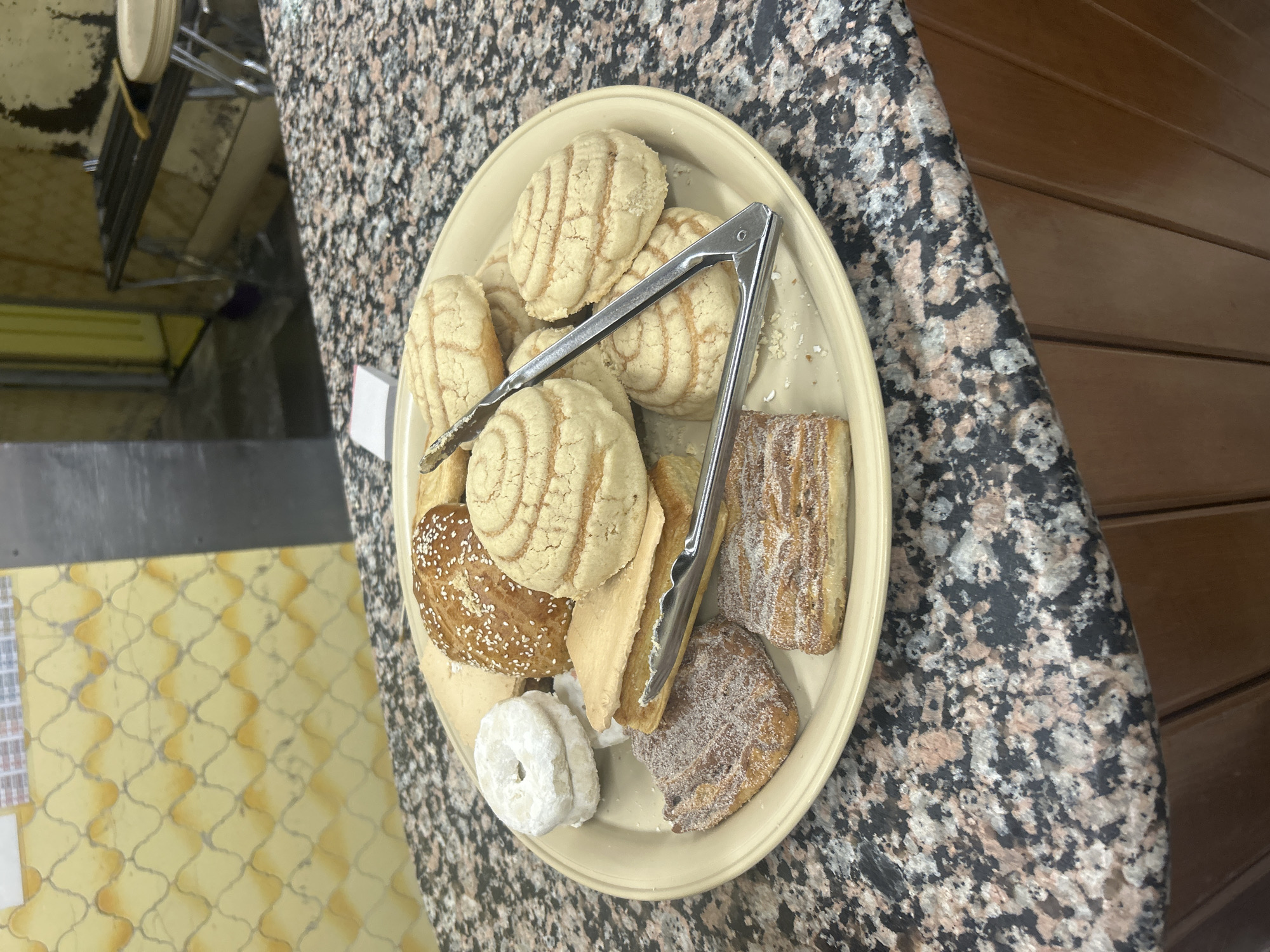 The image shows a plate of assorted Mexican pastries, including conchas, buñuelos, and empanadas, arranged on a beige plate with silver tongs. The plate is placed on a countertop with a speckled pattern. The pastries are dusted with sugar and have various shapes and textures. A portion of the countertop extends to the right, transitioning into wooden panels. In the background, to the left, there's a glimpse of a wall with a checkered tile pattern and a metallic structure, possibly part of a display or serving area. Given the pastries and overall style, it is likely taken in Puebla, Mexico. The scene appears indoors, possibly in a bakery or market setting.