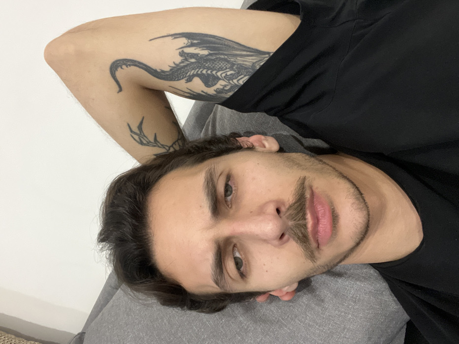 A man is lying down, facing the camera. He has dark hair, fair skin, and a mustache. His left arm is bent behind his head, revealing two tattoos: a dragon and abstract branch-like art. He is wearing a black t-shirt and appears to be resting on a gray pillow. The background is simple, with a white wall visible on the left side.