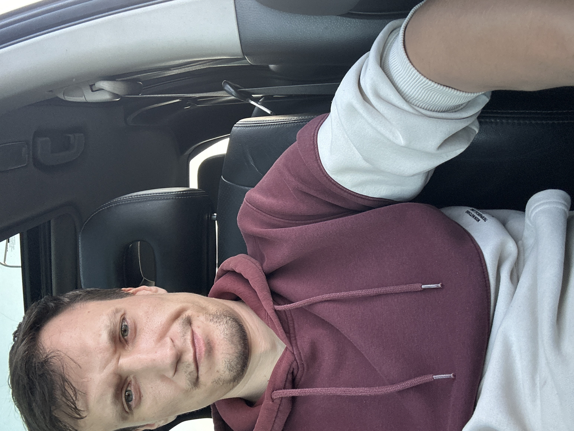 A young adult male with light skin, brown hair, and a short beard is seated in the driver's seat of a car. He is wearing a maroon hoodie over a cream-colored long-sleeved shirt. The interior of the car, including the black leather headrest of the passenger seat and a portion of the dashboard, is visible. The man is looking directly at the camera, and his expression is neutral. The scene appears to be illuminated by natural light, suggesting it is daytime. No specific location within Kamianets-Podilskyi, Ukraine, can be determined from the image.