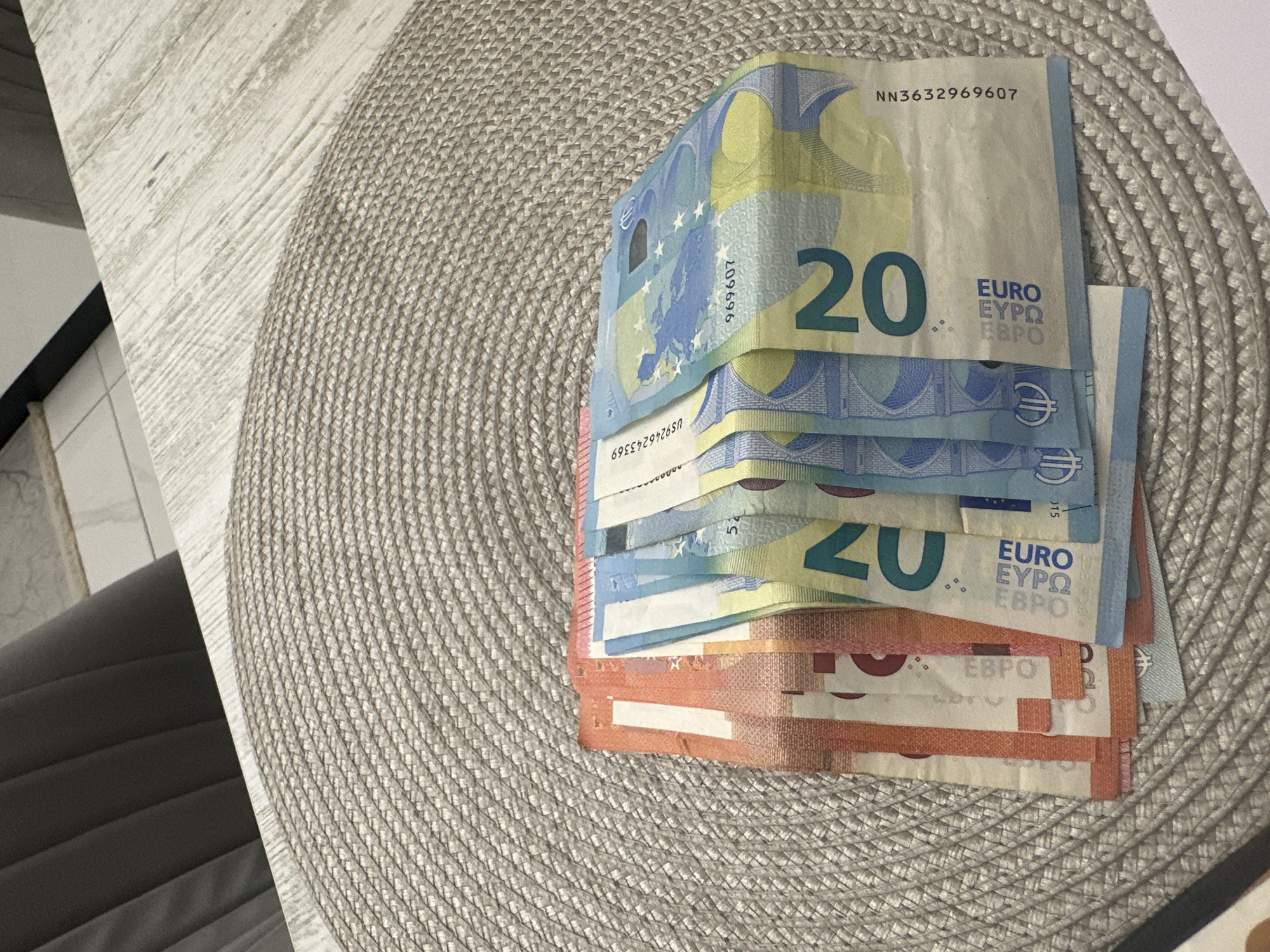 The image captures a static, elevated view of Euro banknotes fanned out on a light beige, circular woven placemat. The placemat rests on a light-colored wooden table featuring a distressed, horizontal plank pattern. On the left, two orange-red 10 Euro banknotes are partially visible. To their right, four blue-green 20 Euro banknotes are arranged, with the number "20" prominently displayed on the exposed portions. Serial numbers visible on the 20 Euro notes include "US216243369" and "NN3632969607".
In the upper left background, a dark grey upholstered chair back with vertical pleats is partially visible. The upper right background reveals a white tiled floor with dark grout lines, adjacent to what appears to be a white wall or baseboard. The scene is illuminated by ambient indoor light, casting soft shadows. No individuals are present, and no actions or interactions are depicted. The specified location of Sofia, Bulgaria, is not visually verifiable within the content of this image.