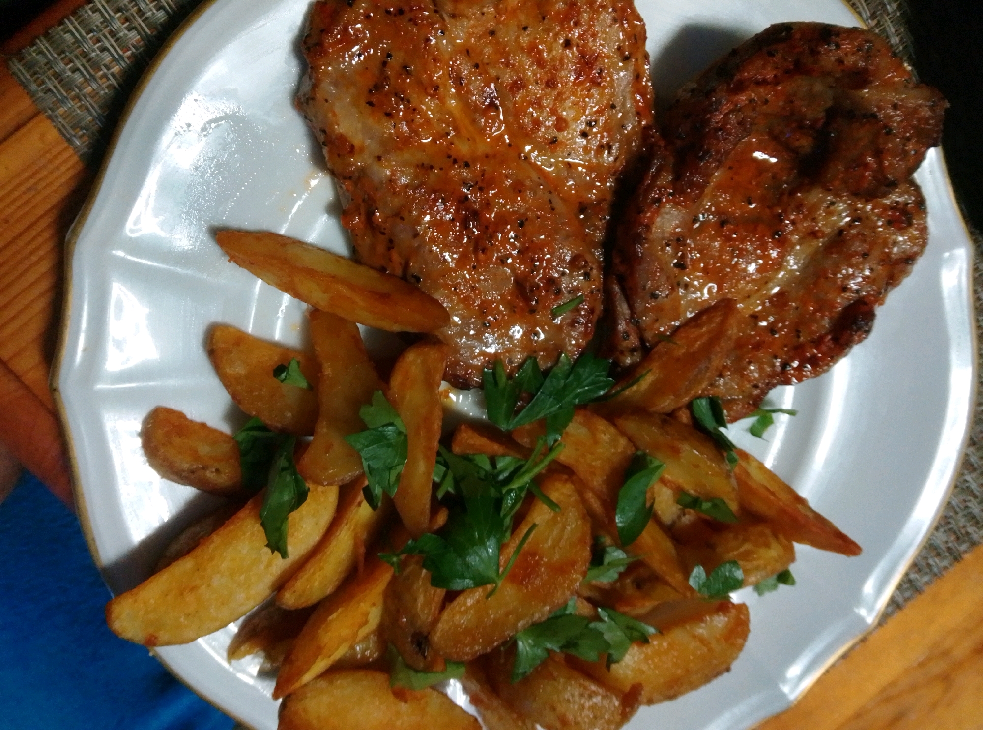 A close-up, top-down shot displays a white, scalloped-edge plate filled with food. Two grilled pork chops, seasoned with black pepper and a glossy sauce, rest on the upper portion of the plate. Scattered around the pork chops are numerous wedge-cut, fried potatoes, some garnished with chopped parsley. The plate sits on a woven placemat with a crisscross pattern in shades of brown and beige. The overall scene is a close-up of a meal, likely prepared in a kitchen. The image does not contain enough information to determine the specific location in the city of Wedel, Germany.