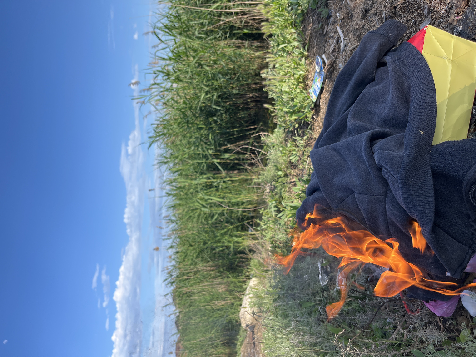 The scene depicts an open area with tall grasses and reeds in the background, under a blue sky with some clouds. A dark-colored hoodie is lying on the ground, near a yellow and red box or package. Flames are emerging from underneath the hoodie, suggesting that something is on fire or has been set on fire. The location is near Kaspiysk, Russia.