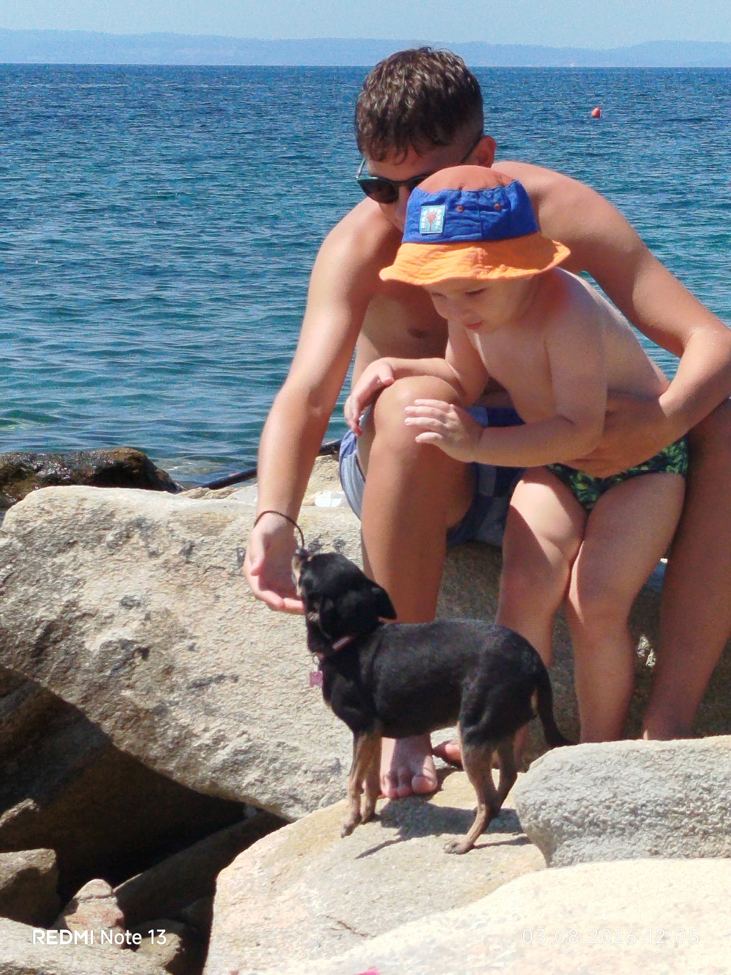 The image shows a man and a young child on a rocky shoreline with the sea in the background. The man is wearing sunglasses and appears to be holding the child. The child is wearing a blue and orange hat and patterned swim trunks. A small black dog is interacting with them, possibly being fed or petted by the man. The water is a deep blue, and the sky is clear and light. The foreground consists of large, textured rocks. The scene appears to be outdoors, likely at a beach or coastal area. The "REDMI Note 13" watermark suggests the image was taken with a Redmi Note 13 phone. The "03.08.2026 12:05" watermark indicates the date and time the photo was taken. The location is Sithonia Municipal Unit, Greece.