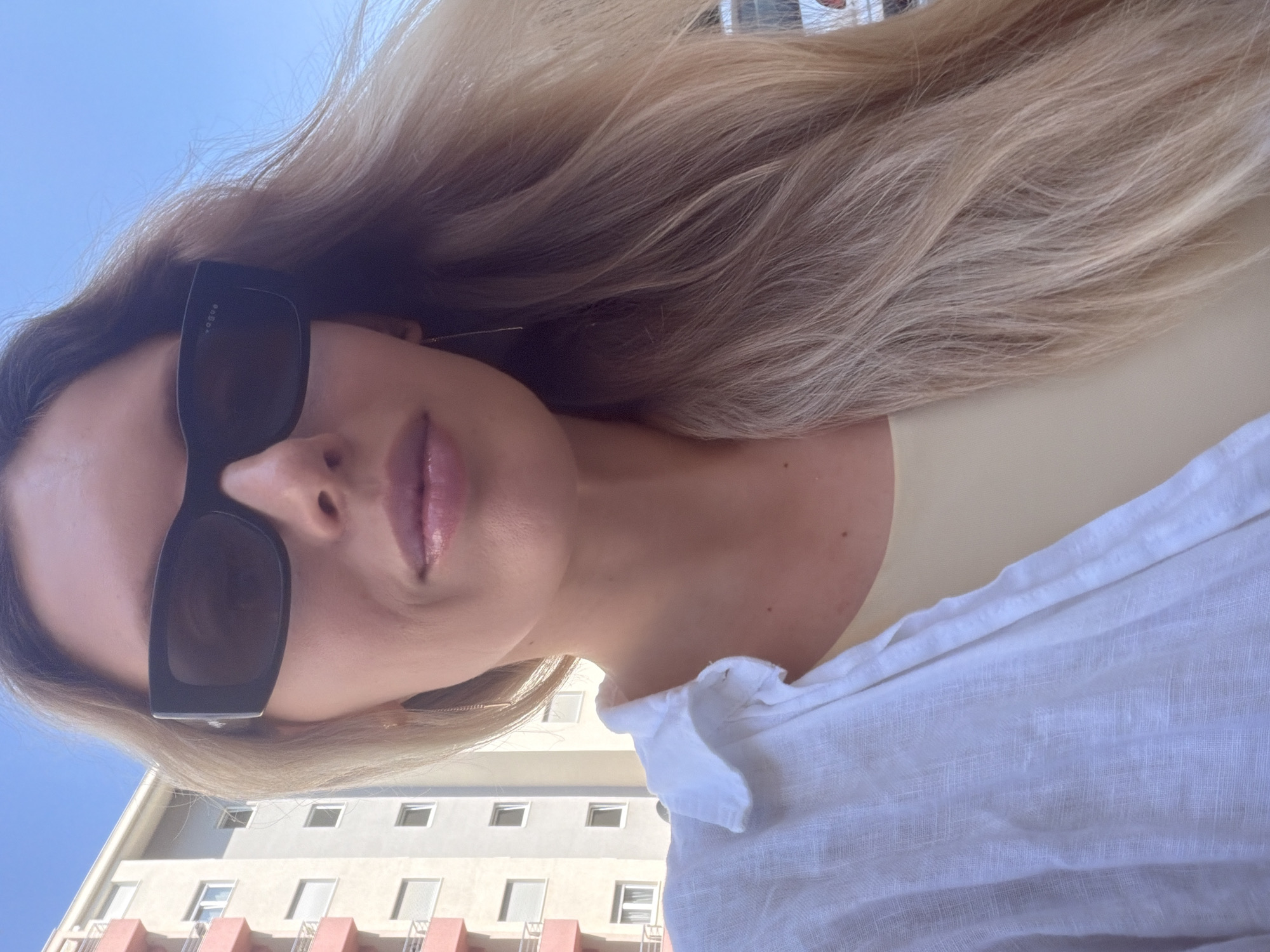 A woman is taking a selfie outdoors in front of a building. She has long blonde hair and is wearing black sunglasses and a white shirt over a light yellow top. The background shows a modern building with balconies and windows, and a clear blue sky. The woman's face is partially obscured by her sunglasses. The lighting suggests it is daytime.