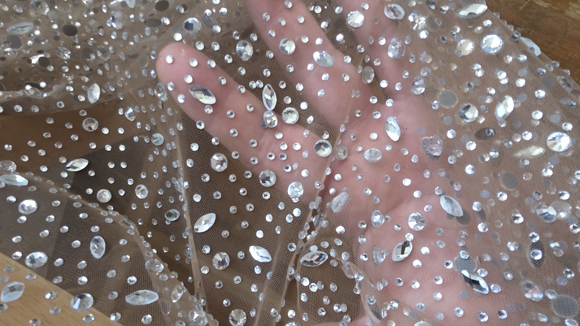 The image displays a close-up, high-angle shot of a sheer, light-colored mesh fabric lavishly adorned with numerous clear, sparkling rhinestones and crystals. A human hand is visible beneath the translucent material, its fingers and palm slightly blurred, emphasizing the fabric's sheerness.
The fabric appears to be a fine net or tulle, exhibiting a warm, nude, or very light brown tone. It is densely embellished with reflective elements that catch the light, creating a glittering effect. These embellishments vary in size and shape; small, round rhinestones are scattered across the surface, interspersed with larger, faceted oval and marquise-shaped crystals. The clarity and varying cuts of the stones contribute to their brilliance.
The hand underneath the fabric is positioned to showcase the material, with the individual digits and the palm clearly outlined through the embellished mesh. The texture of the fabric and the distinct attachment of each rhinestone are discernible upon closer inspection.
The background is softly out of focus, suggesting an indoor setting. Hints of a light brown, possibly wooden, surface are visible beneath the fabric, particularly on the left side of the main image. There are no specific cues within the image to indicate the location as Blagoevgrad, Bulgaria. No text is visible, and no particular activity is taking place beyond the presentation of the sparkling, sheer textile. The overall impression is one of delicate opulence and shimmer.
