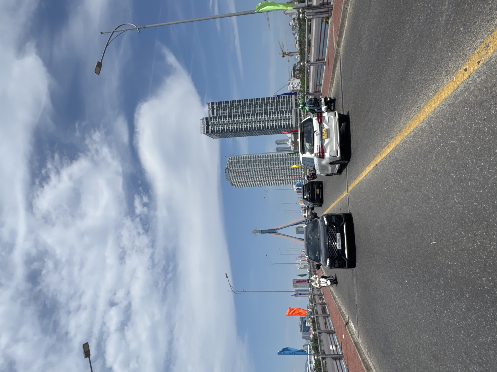 The image shows a view from a bridge in Đà Nẵng, Vietnam. On the bridge, two cars and a scooter are traveling along the roadway. A solid yellow line marks a traffic lane. In the background are skyscrapers and construction cranes. Flags line the edge of the bridge. Streetlights are visible along the bridge. The sky is blue with scattered clouds.
