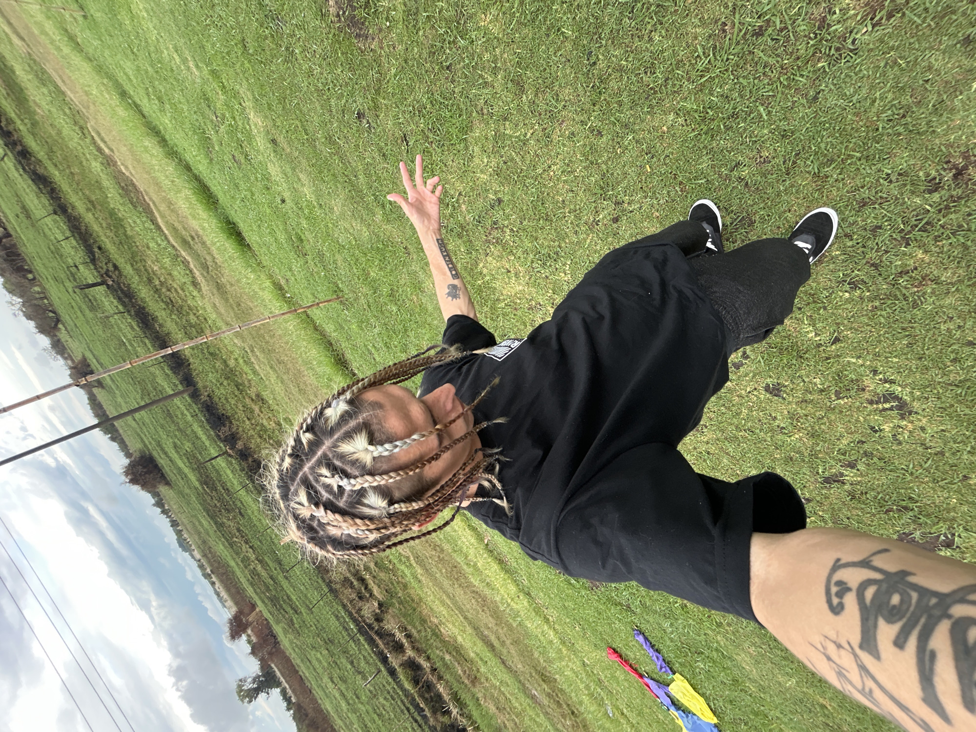 The image shows a person lying on green grass, presumably in Dolores, Argentina. The person is facing the camera and has light-colored braided hair. They are wearing a black t-shirt and dark pants, along with black shoes. Their right arm is outstretched, making a peace sign, and has a tattoo on the forearm. The left arm is also visible and has tattoos. In the bottom right corner, a small colorful object resembling miniature umbrellas is visible. In the background, there's a grassy field extending towards a line of trees and a partly cloudy sky. Utility poles and wires are also visible in the background.