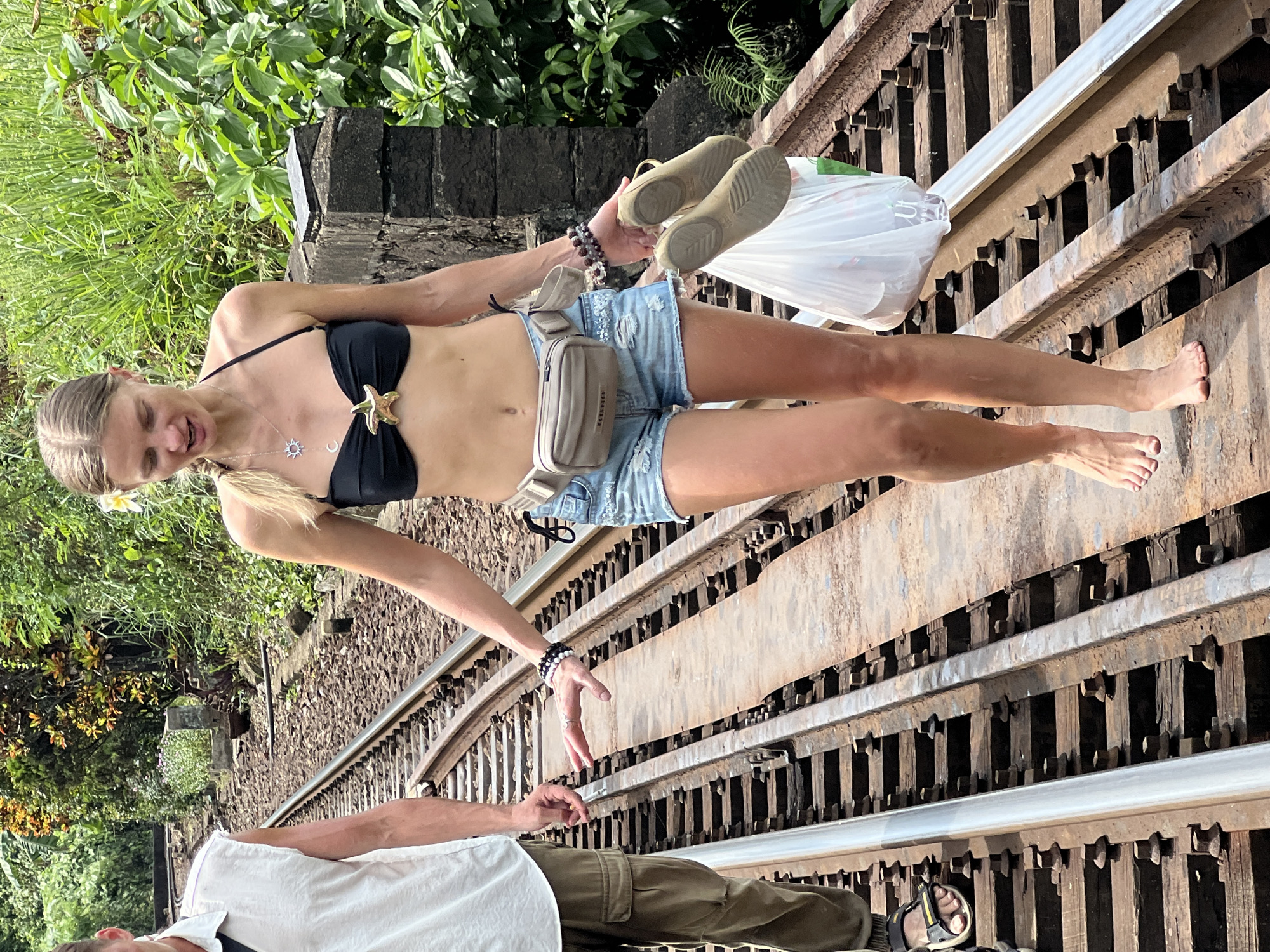 Two individuals are walking on a railway track, likely in or near the city of Huế, Vietnam. The person on the right is a woman with blonde hair, wearing a black bikini top, distressed denim shorts, and a beige fanny pack. She is barefoot and carrying a white plastic bag with a pair of beige sandals. She has several bracelets on her left wrist and a necklace with a sun and crescent moon charm. The person on the left, partially visible, appears to be a man wearing a sleeveless white shirt and olive-colored cargo pants. He is wearing sandals on his left foot. The railway tracks are made of metal rails and wooden sleepers, surrounded by lush green vegetation and trees. The scene appears to be outdoors, under natural daylight.