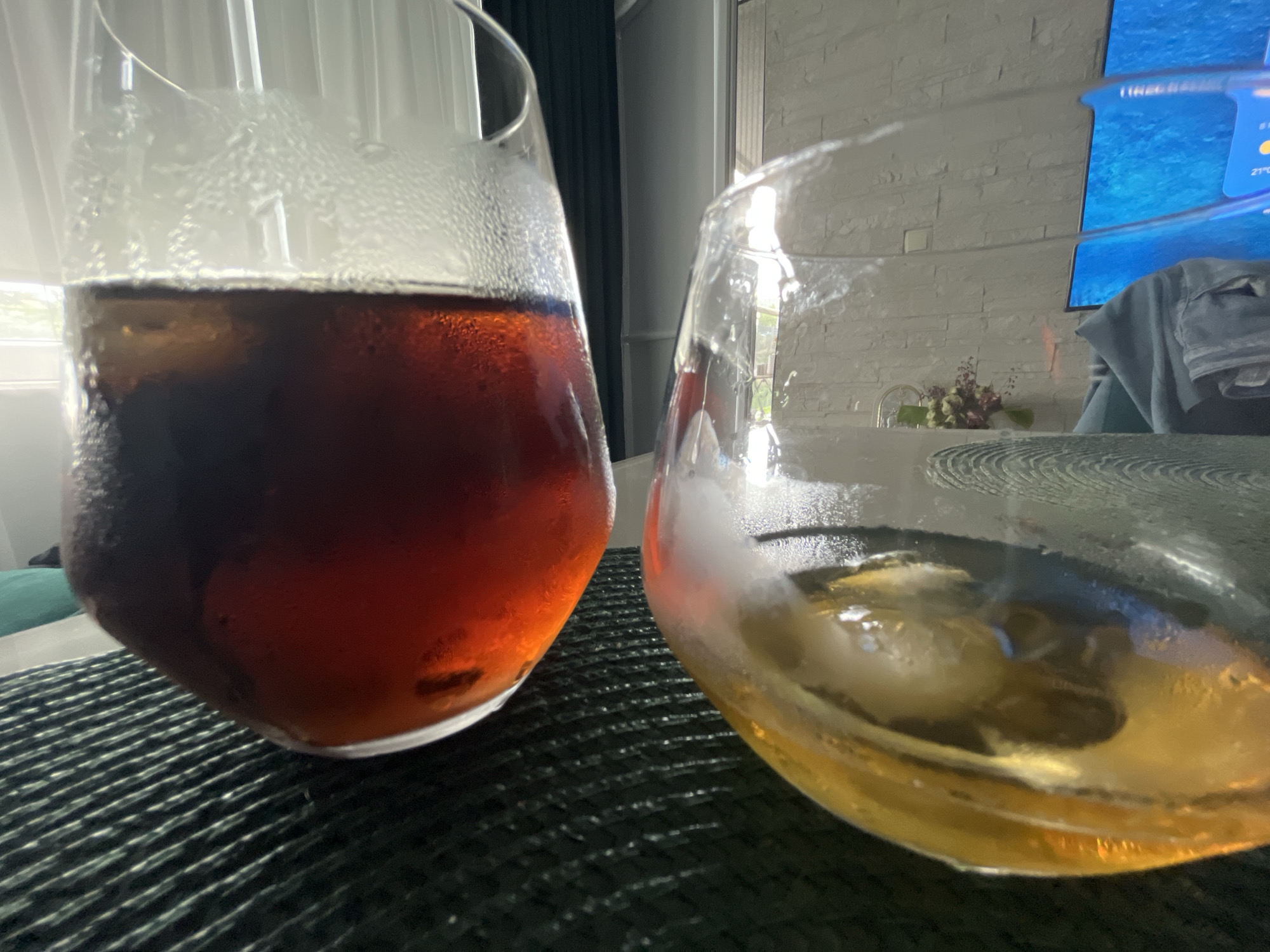 Two wine glasses are on a dark placemat. The glass on the left contains a dark brown liquid with ice, and the glass on the right contains a light brown liquid with ice. The glasses are sweating, indicating they are cold. In the background, a television is on, displaying a blue screen with some text. To the left of the television, a window with white blinds is visible. A blue curtain is partially visible to the left of the window. The scene appears to be indoors, possibly in a residential setting, with a modern aesthetic. The location in Varna, Bulgaria, is not discernible from the image.