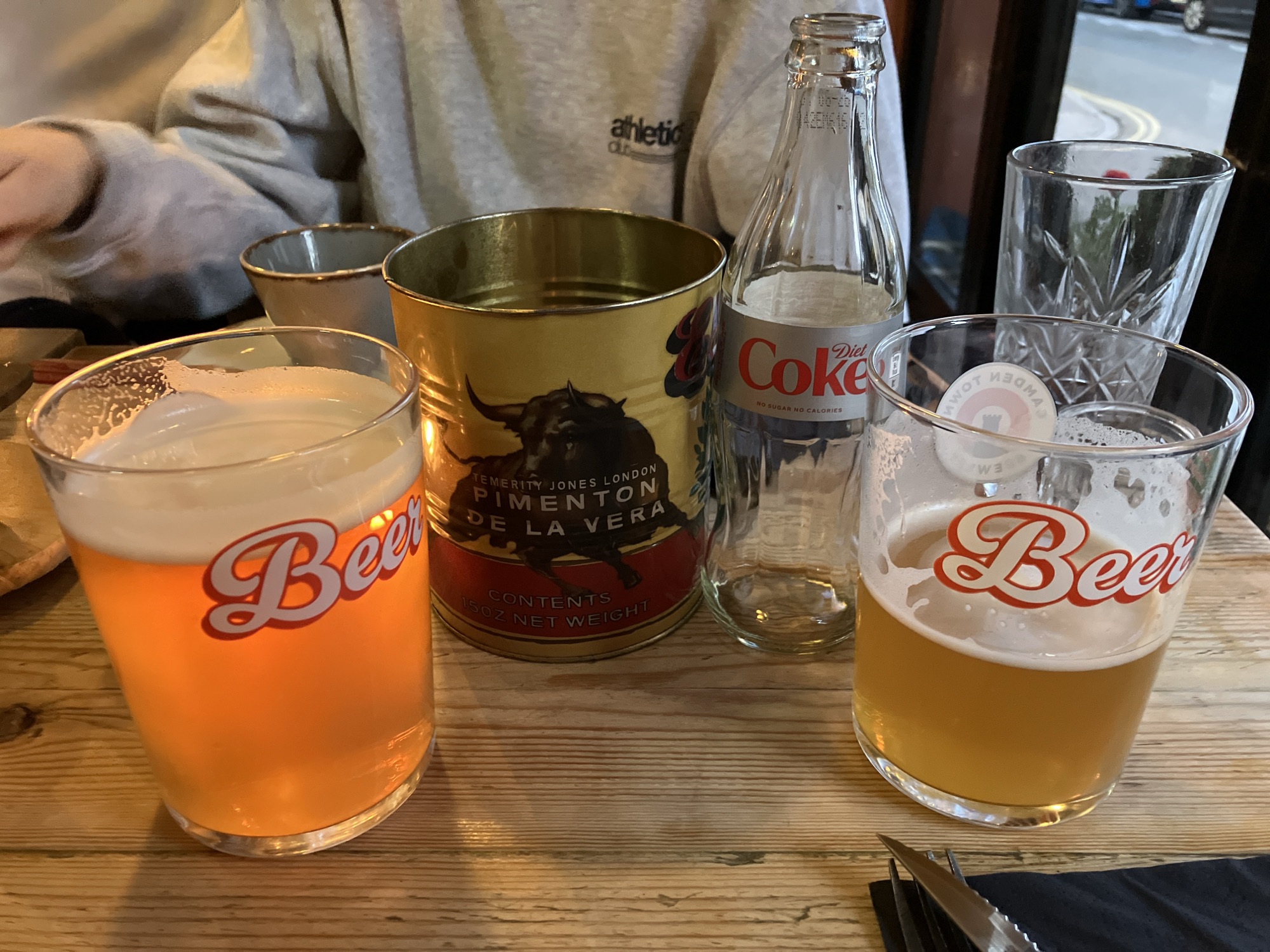 A table arrangement features two beer glasses with "Beer" emblazoned on them, alongside a tin of "TEMERITY JONES LONDON PIMENTON DE LA VERA," a glass bottle of Diet Coke, and two additional glasses. The setting seems to be indoors, with a person in an "athletic" sweatshirt visible in the background. The scene is set on a wooden surface.