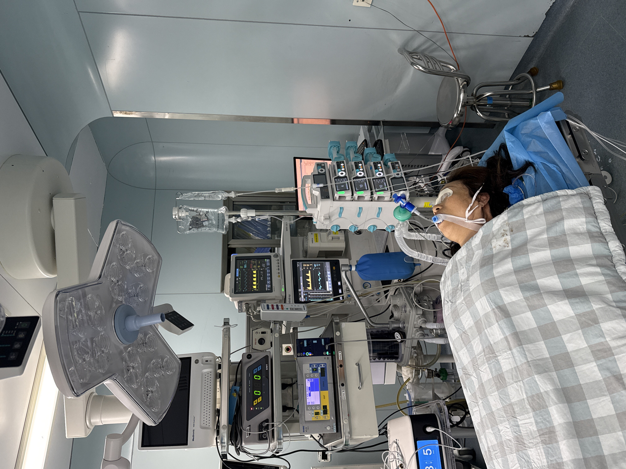 The image shows a person lying in what appears to be an operating room or intensive care unit, surrounded by medical equipment. The person is covered with a checkered blanket and has a mask over their nose and mouth, along with protective pads over their eyes. Various monitors and machines display vital signs and settings. A surgical lamp is visible in the foreground, and IV bags hang nearby. The room has light blue walls, suggesting a sterile environment.
The exact location within Yuzhong District, China, cannot be determined solely from the image.