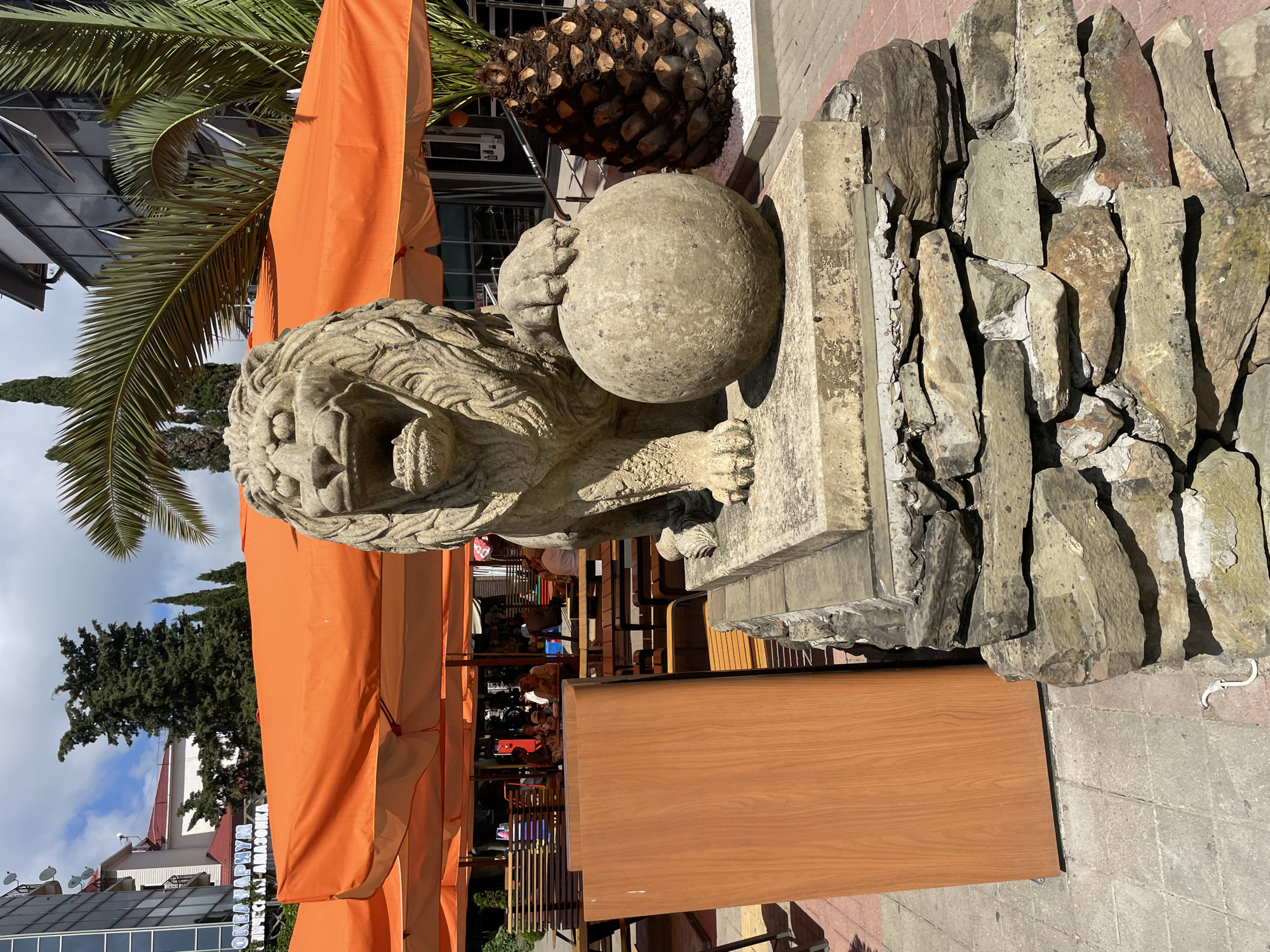 This is an outdoor photograph of a stone lion statue in Sochi, Russia, near the coast. The statue is centrally located in the foreground, depicting a lion with its mouth open, resting its paw on a sphere. The statue is placed on a multi-tiered base, with the lower section made of rough, stacked stones and the upper section a lighter-colored stone platform. Behind the statue, several orange umbrellas cover an outdoor seating area of a cafe or restaurant. The umbrellas are partially obscuring tables and chairs, and in the background, people are seated and interacting. To the right of the statue, a palm tree with a distinctive textured trunk stands in a bed of white gravel. Further in the background, modern buildings with glass facades and balconies are visible, along with other trees, including cypress trees and palm trees. The sky is partly cloudy, with clear blue sky visible. The ground is paved with interlocking concrete tiles. The overall scene suggests a sunny day in a tourist-oriented area.
