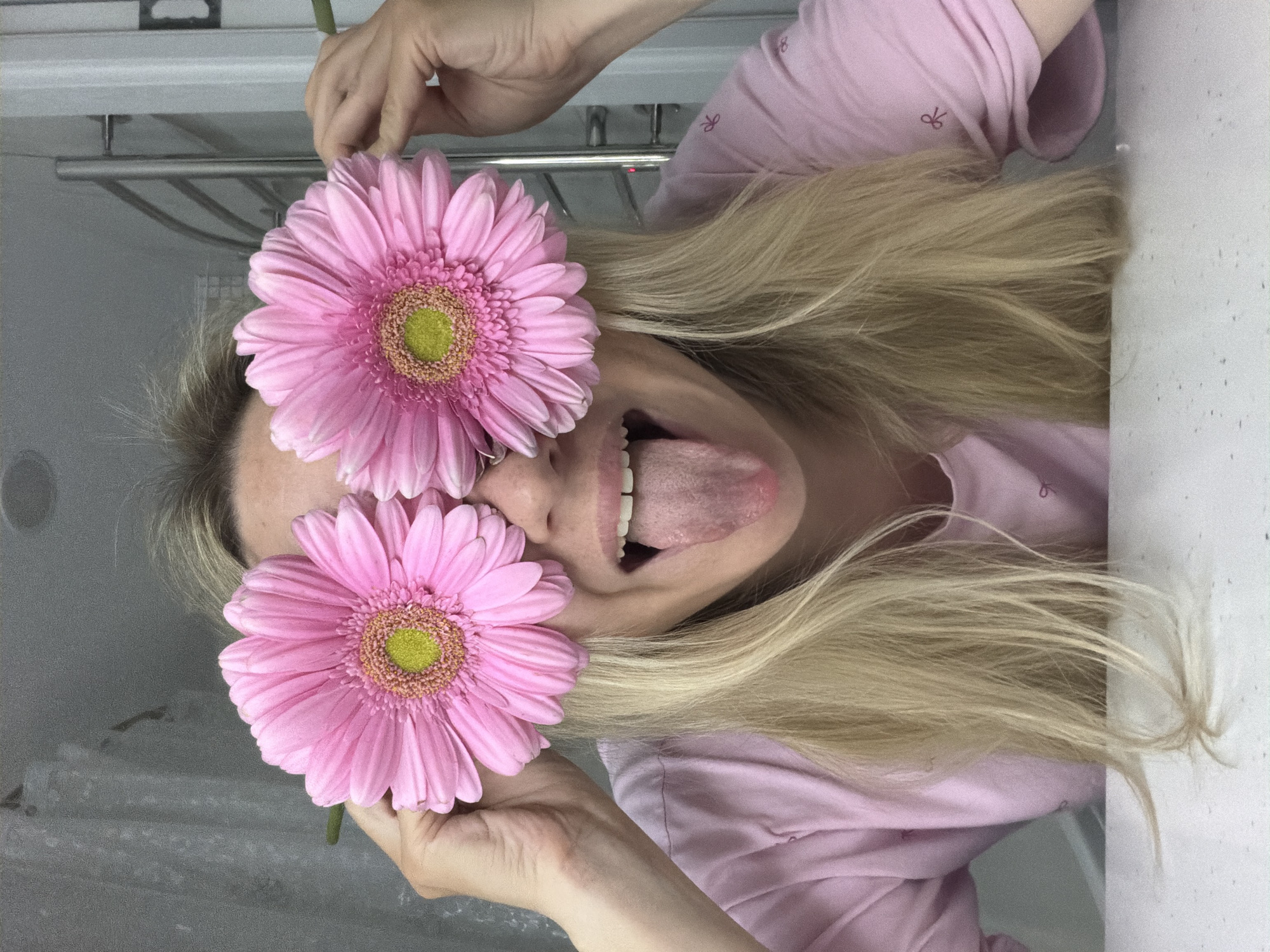 A person with long blonde hair, wearing a pink shirt, is holding two pink gerbera daisies over their eyes and sticking their tongue out. The background shows a bathroom with a shower rack visible. There is no indication of Votkinsk, Russia.