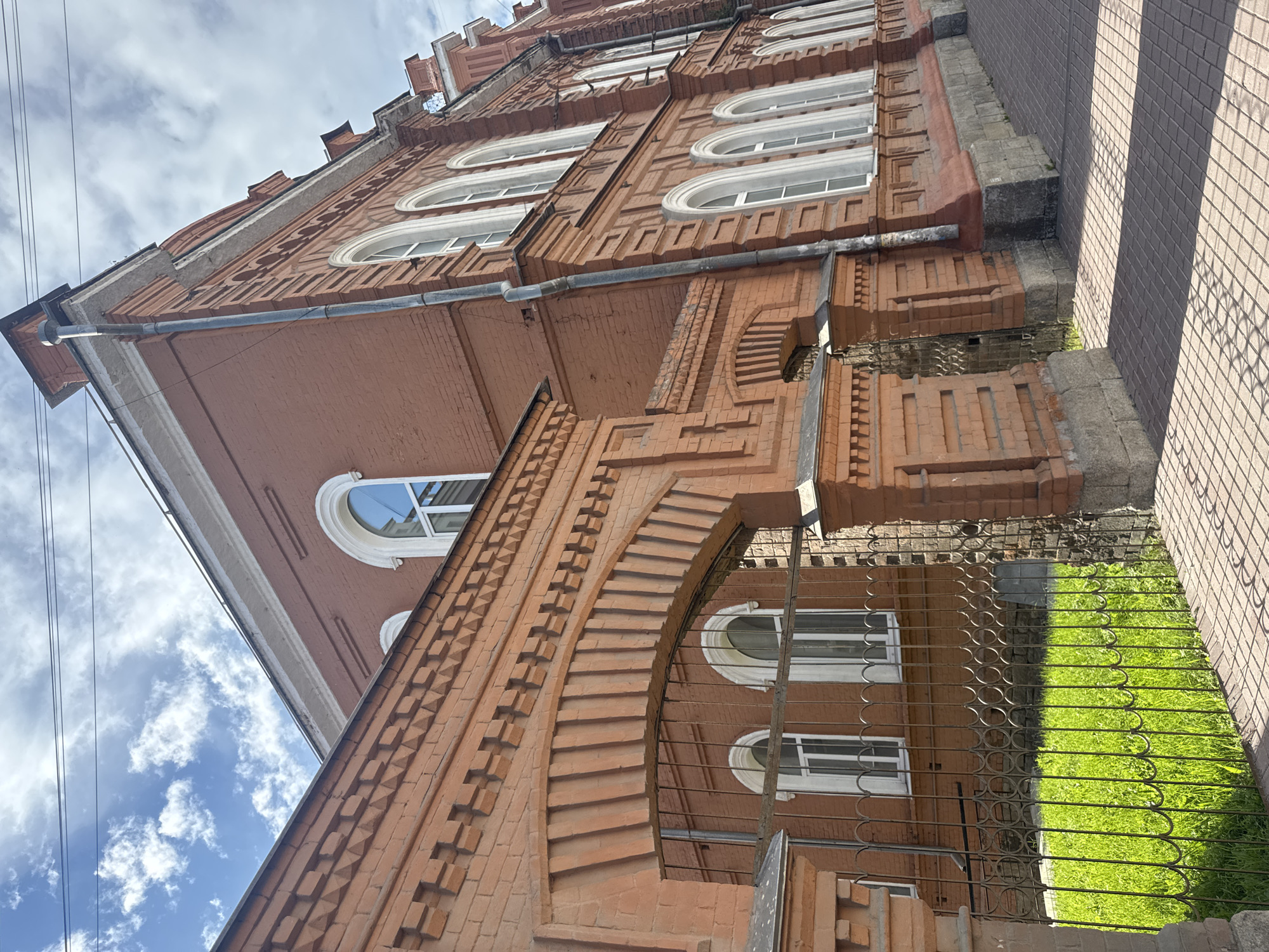 This is a low-angle, outdoor, full-shot photograph of a multi-story brick building. The building appears to be an older architectural style, with decorative brickwork and arched windows. The brick is predominantly a reddish-brown color. Several windows are visible, each with a white frame and what appears to be clear glass. Some of the windows are rounded at the top, in an arched style. The building has a tiled roof, and a drainpipe runs along the side.
In the foreground, a decorative metal fence with a repeating circular pattern encloses a patch of bright green grass. Beyond the fence, the lower part of the building features more intricate brickwork in an arched doorway or entryway.
The sky above is a mix of blue and white clouds. The lighting suggests it is daytime, likely late morning or early afternoon, given the shadows cast by the building on the adjacent tiled walkway. The photograph is taken at a tilt, with the building receding upwards and to the right of the frame. No people are visible in the image. The setting appears to be urban, possibly a residential or institutional building in Yekaterinburg, Russia. There is no visible text in the image.