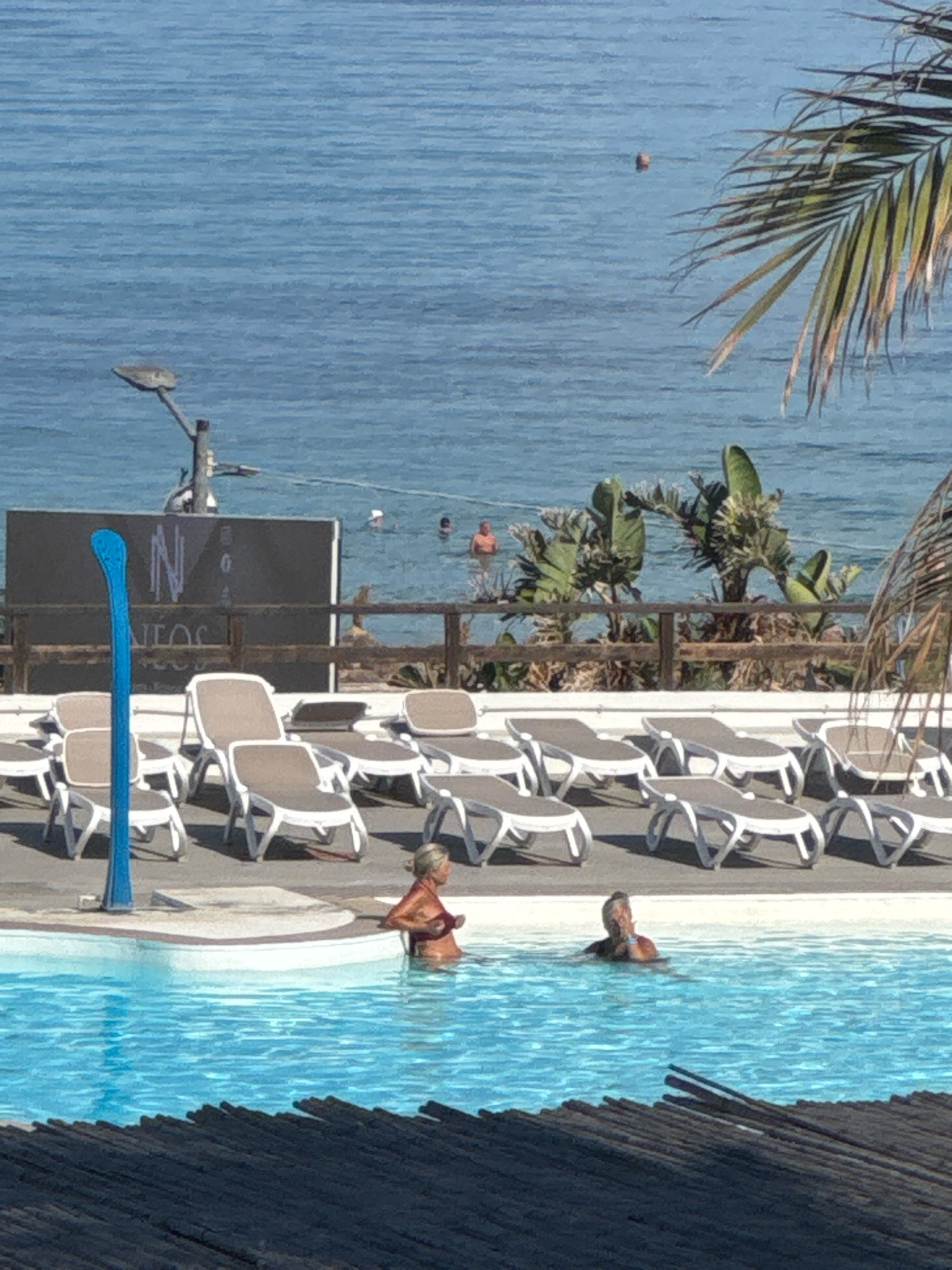 This is a photograph taken at a resort in Capaci, Italy, overlooking the Tyrrhenian Sea. In the foreground, a bright blue swimming pool dominates the lower half of the image. Two people are in the pool: a woman on the left, facing away from the camera and towards the sea, wearing a red bikini, and a man on the right, partially submerged, looking towards the camera.
Behind the pool, a row of white and beige lounge chairs are arranged on a paved surface. Further back, a wooden fence separates the resort area from the beach and the sea. A dark sign with white lettering, which appears to read "NEOS," is mounted on a pole behind the fence. In the background, the calm, blue water of the sea stretches to the horizon. A few people are visible in the sea, and a lone object, possibly a buoy, floats on the surface. Palm trees and other green foliage are scattered along the coastline. The sky is a clear, bright blue.