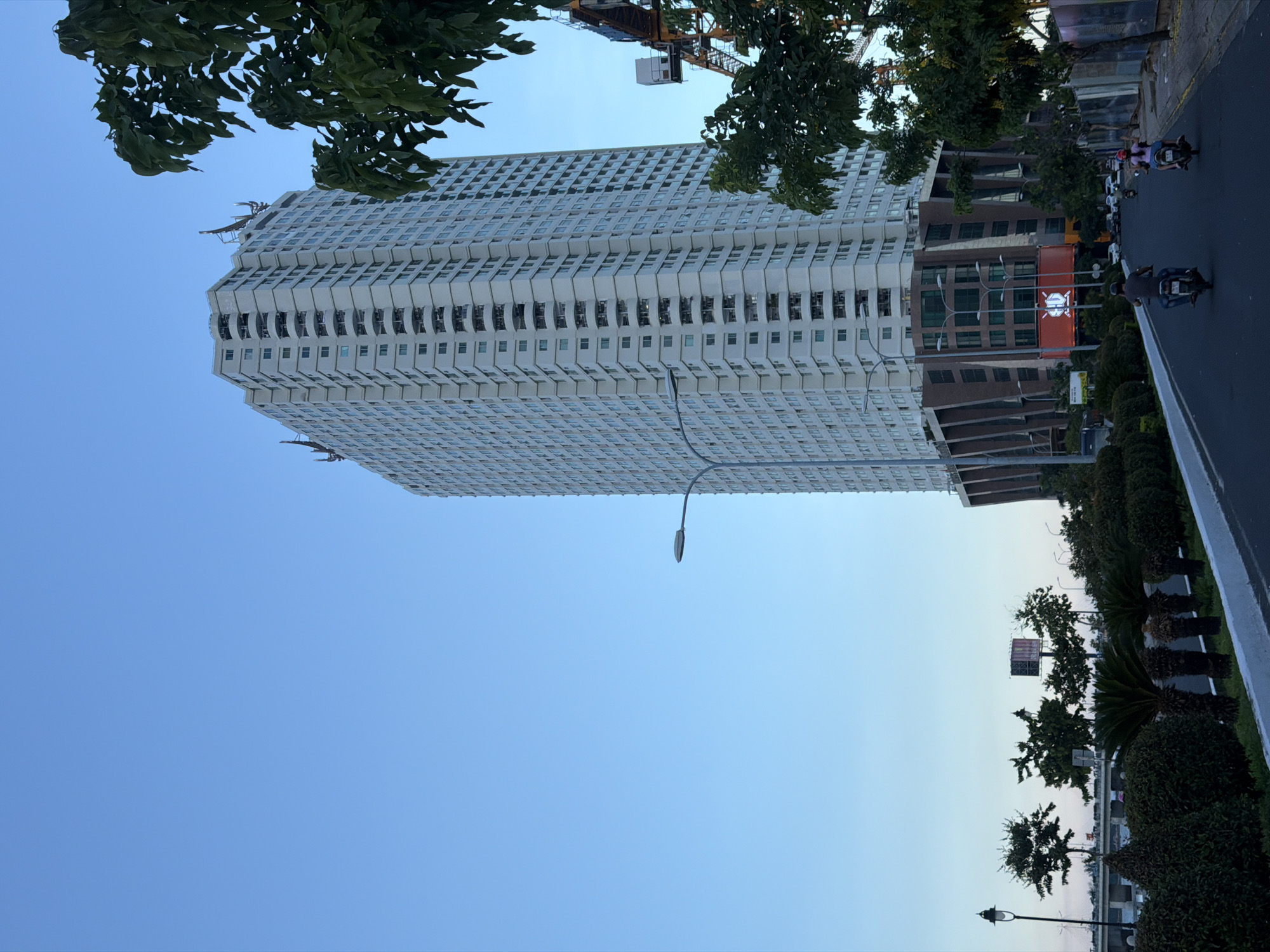 The image shows a modern high-rise apartment building in Nha Trang Ward, Vietnam, on a clear day with a bright blue sky. The building is tall and rectangular, with many floors and windows. It has a white exterior and a distinctive design on its lower levels with brown, angular elements and large windows. In front of the building, there is a street with a few motorbikes carrying people, suggesting traffic. Trees and lush green bushes line the street and the sidewalk. In the background, there are more trees, a distant bridge, and construction equipment, including a crane. The overall scene depicts an urban environment with a blend of residential buildings, infrastructure, and greenery.