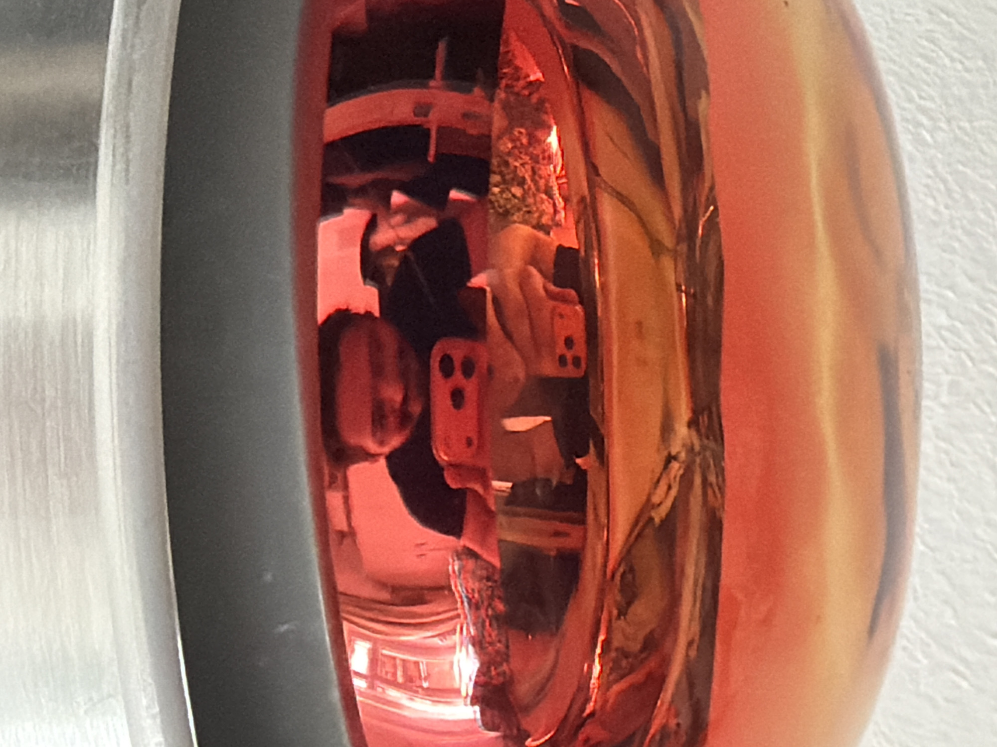 The image shows a close-up reflection in a metallic, reddish-orange, possibly spherical object. The reflection contains a person holding a mobile phone (likely an iPhone based on the square camera module with three lenses). The person is partially obscured by the object reflecting them. Parts of an interior space, possibly a room, are visible in the reflection. The background is out of focus and appears to be a white textured surface. The left edge of the image is a grey metallic object. It is not possible to determine the location in Belgrade, Serbia from this image alone.