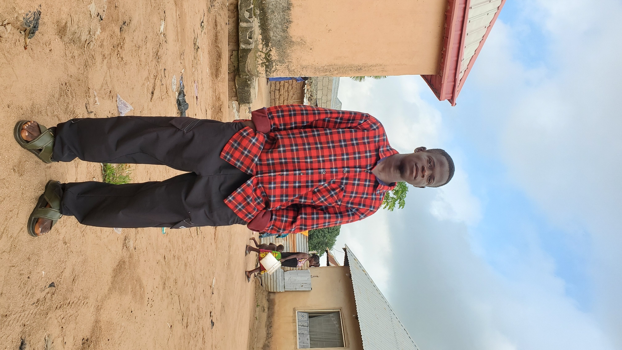 A young Black male stands facing forward in a village setting. He is wearing a red and black plaid long-sleeved shirt with the sleeves rolled up, black cargo pants, and green open-toed sandals. His hands are in his pockets. Behind him, to the left, is a tan building with a red roof. To the right, another building with a silver corrugated metal roof is visible. In the background, two other individuals, a woman and a child, are walking. The ground is sandy dirt, with some scattered debris. The sky is partly cloudy.
