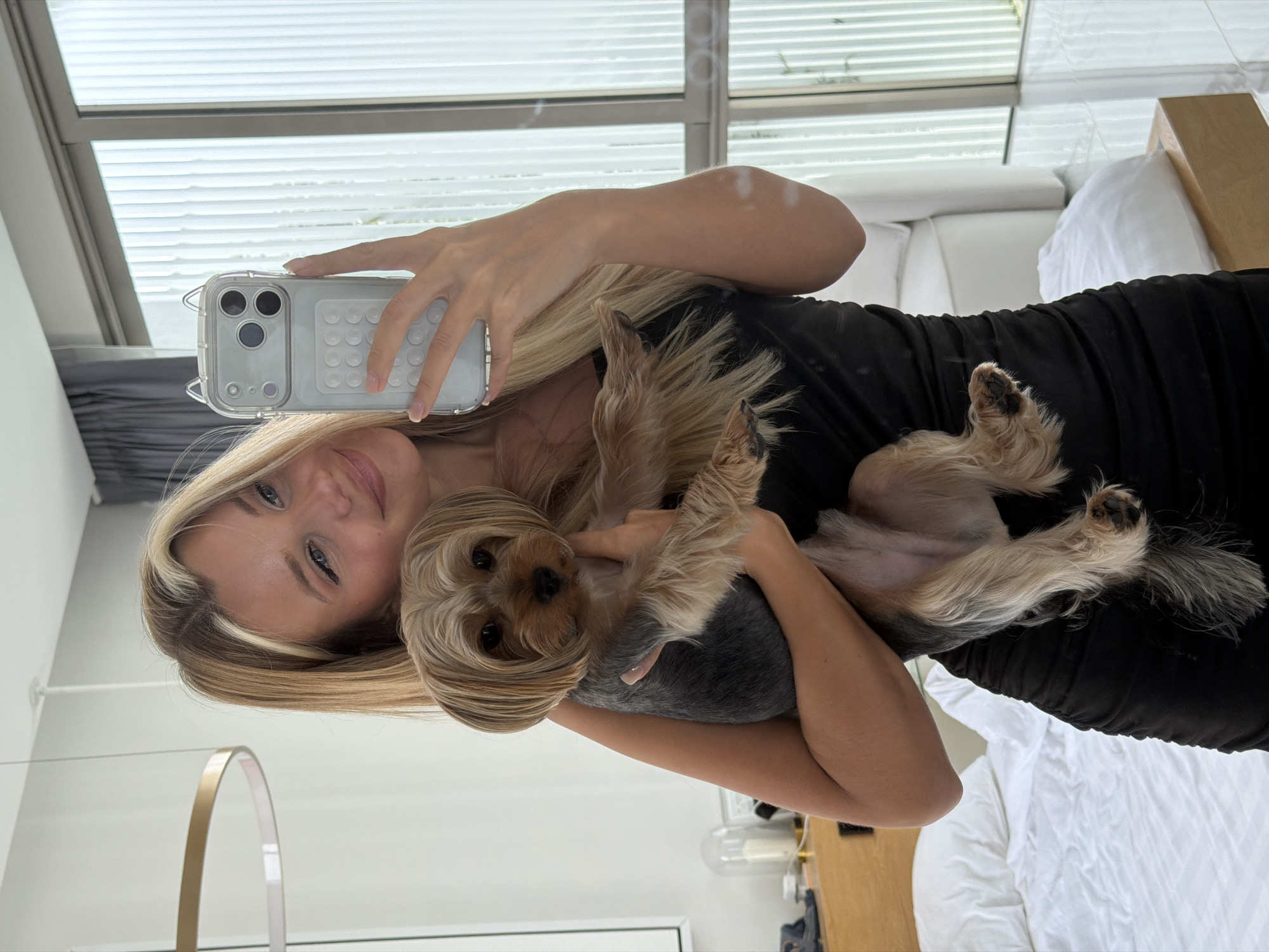 A blonde woman in a black dress is taking a mirror selfie with her yorkshire terrier. She is holding the dog in her arms and looking at the camera with a smile. The dog is also looking at the camera. The background is a bedroom with a white bed and a window. The location is a modern bedroom in Thep Krasatti, Thailand.