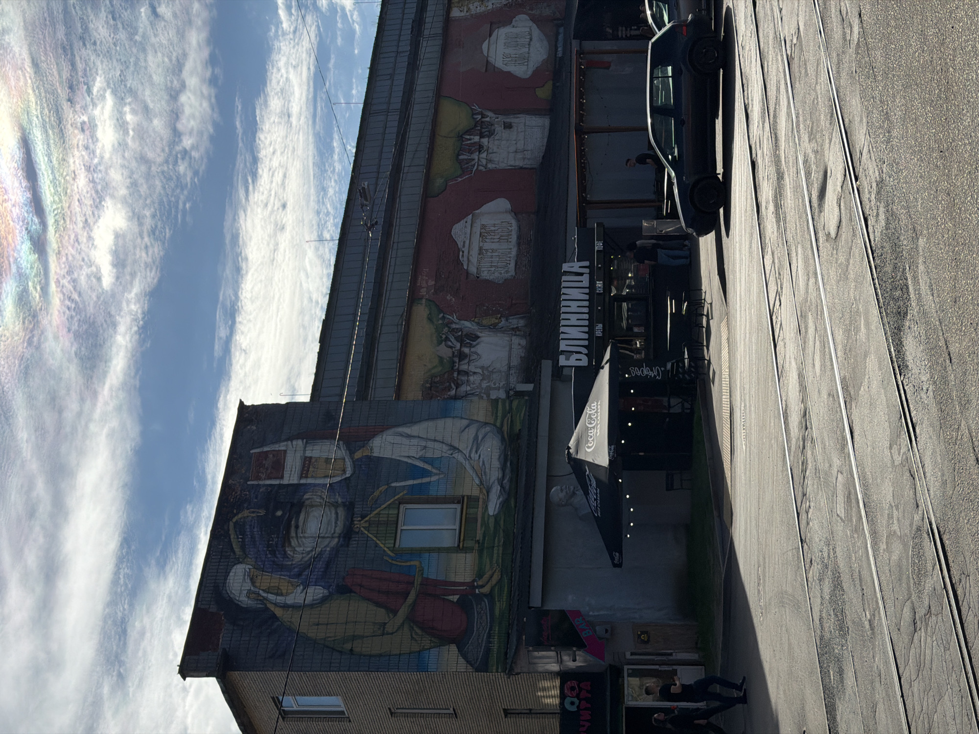 The image depicts a street scene in Minsk, Belarus, featuring a building with prominent murals. The building's facade is decorated with large, colorful artwork. On the left side of the building, a mural shows two figures, one in red trousers and the other in white, sitting near a window. To the right, another mural displays abstract elements and what appears to be text in Cyrillic. Below the murals, a covered outdoor seating area is visible, with a sign displaying "БЛИННИЦА" (Blinnitsa, meaning "pancake maker" or "pancakes"). A Coca-Cola umbrella is also present, as is a statue of Lenin. Several people are seen walking on the sidewalk and near the building. In the foreground, tram tracks run across the asphalt street, and a dark-colored car is parked on the right side of the road. The sky above is blue with scattered clouds, some of which exhibit iridescent colors, suggesting a phenomenon like a cloud iridescence. The overall impression is a public space with artistic elements and commercial activity.