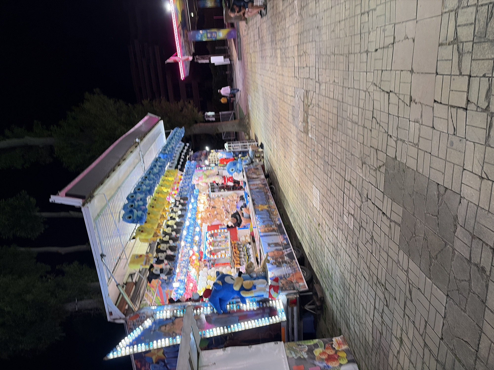 A nighttime scene at what appears to be an amusement park or carnival in Unknown, Unknown. A game booth, decorated with colorful plush toys including Sonic the Hedgehog and what looks like Stitch, is prominently featured on the left. The booth is illuminated with bright lights, and various prizes are displayed. To the right, a paved pathway leads towards other attractions, also lit up with neon signs. A few people are visible in the background, walking or sitting, some appearing to be staff members. The overall atmosphere suggests a lively evening event.