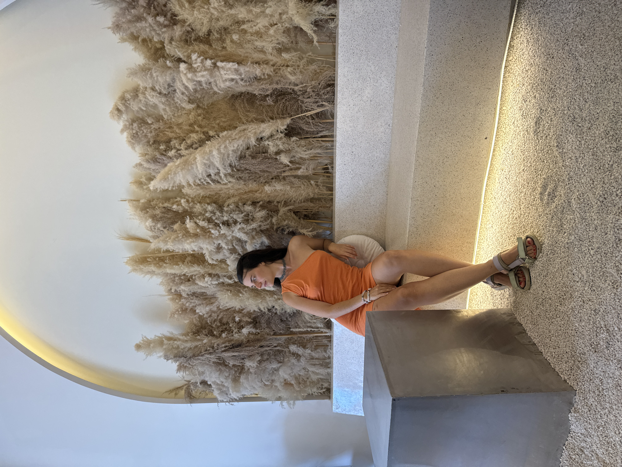 A woman is depicted lounging on a low, light-colored bench or platform in a contemporary indoor setting. She is wearing an orange, off-the-shoulder top or dress and light-colored sandals, with her legs crossed at the ankles. Her dark hair is swept back, and she looks to the side with a calm, neutral expression.
The dominant feature of the background is a large wall densely adorned with dried pampas grass, creating a soft, textured, and natural aesthetic. The architecture includes a curved, smooth white wall with a strip of warm, yellow light embedded along its edge, providing indirect illumination. The floor visible to the right of the woman is covered in small, light-colored pebbles or a textured aggregate material. Other surfaces appear to be light-toned stone or concrete, contributing to a minimalist yet inviting atmosphere.
The lighting is warm and ambient, suggesting an interior space designed for relaxation, possibly a cafe, restaurant, or hotel lobby. The overall scene conveys tranquility and modern elegance. No visible text is present in the image. The setting is located in Phu Thuy Ward, Vietnam.