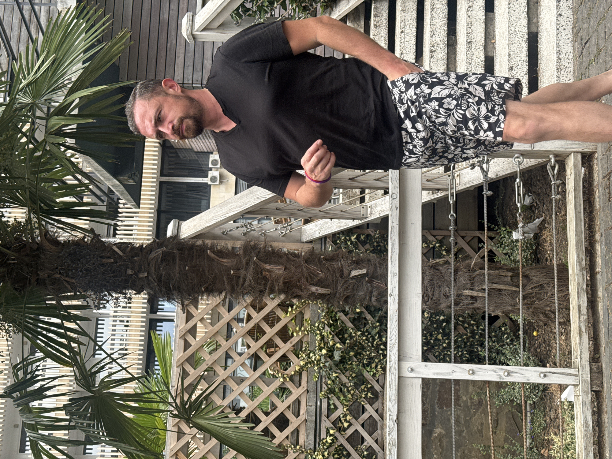 A man with a beard stands outdoors in front of a palm tree and a wooden structure with stairs. He is wearing a black v-neck t-shirt and black and white floral patterned shorts. His right hand is in his pocket, and his left hand is slightly raised. The background shows a building with white vertical slats and windows. There is a wooden lattice fence with ivy growing on it. The ground is paved with cobblestones and dirt. The image appears to be taken in a warm climate. The presence of a palm tree, the architectural style of the building, and the overall ambiance suggest a location in a city like Yalta, Ukraine, known for its seaside resorts and subtropical vegetation.
