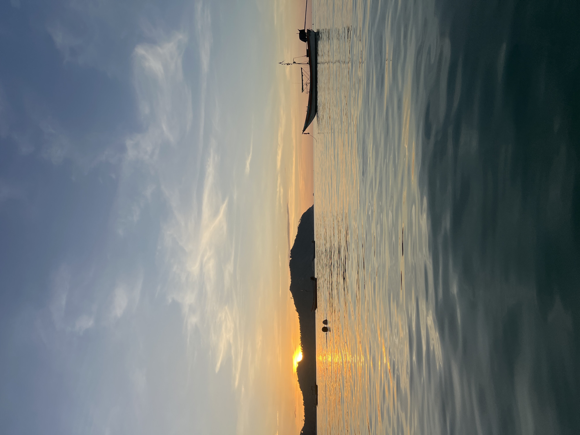 This image depicts a serene sunset scene at sea, likely near ตำบลเกาะพะงัน, Thailand. The sun, partially obscured by a distant, dark-silhouetted island, casts a warm orange glow across the sky and reflects vibrantly on the rippling water in the foreground. Wispy white clouds are scattered across the blue sky.
Several boats are visible in the middle ground. One prominent longtail boat, silhouetted against the horizon, is moored on the right side of the frame. Its anchor rope is visible trailing into the water. To the left, closer to the island, are two smaller boats, also appearing to be at anchor. In the water between these boats, two buoys are visible, floating on the surface. The water transitions from a bright, reflective orange near the sunset to a darker, deep blue-green towards the bottom of the frame, with gentle waves creating subtle patterns on its surface. The overall impression is one of tranquility and natural beauty at the close of day.