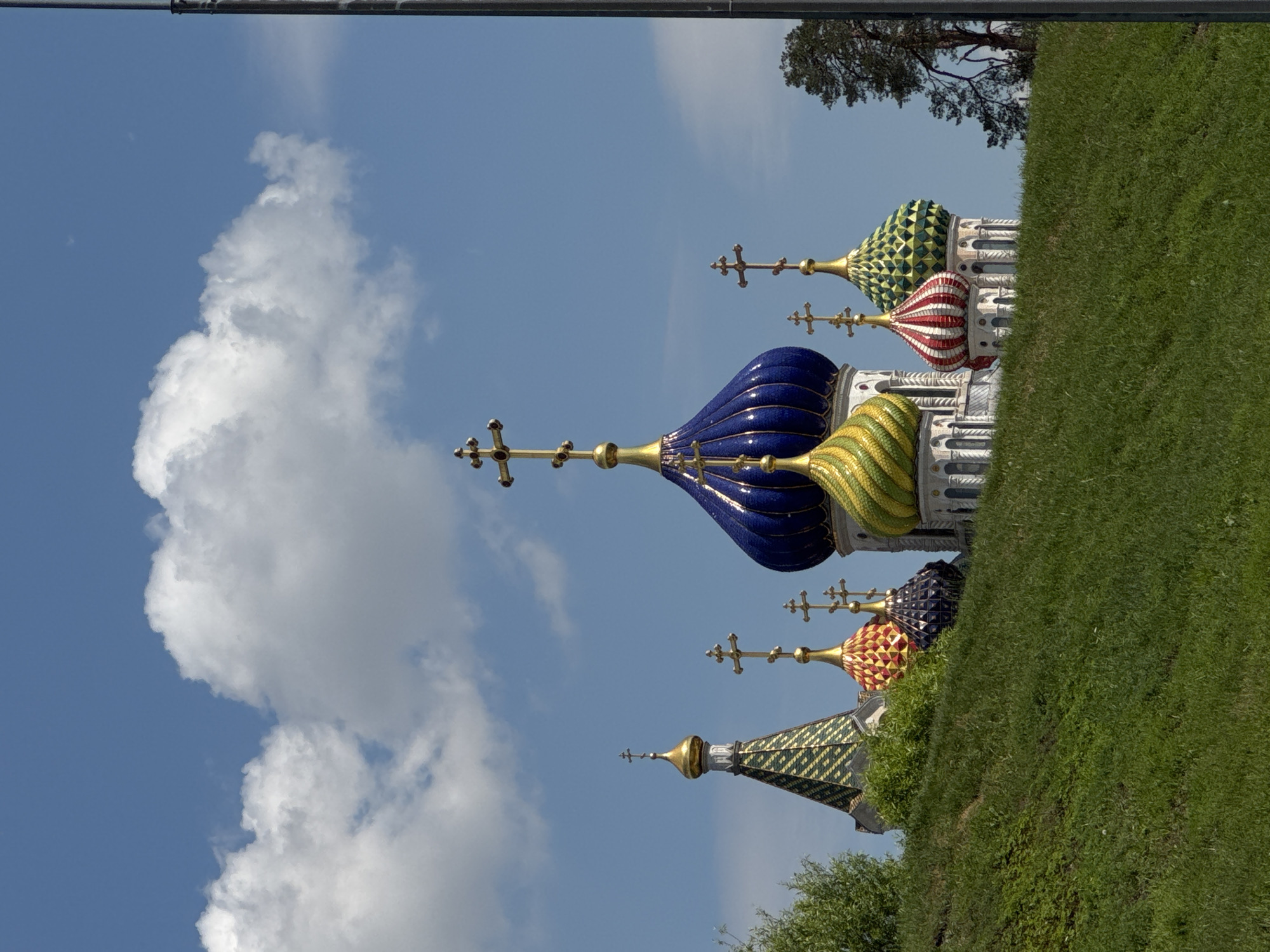 This brightly lit, full-shot photograph features a collection of colorful, ornate onion domes adorned with golden Orthodox crosses, characteristic of Russian architecture. The domes emerge from behind a vibrant green, grassy slope that occupies the lower portion of the frame.
In the center, a large, prominent blue dome with a golden cross draws attention. To its right, a smaller golden-yellow spiraled dome is visible. Further right, other domes include a striking red and white striped one, a green patterned one, a dark blue/purple patterned dome, and a reddish/orange patterned dome. On the left side, a distinct green and gold patterned dome with a single cross is also present. Beneath the domes, intricate white architectural elements of the church structures can be seen.
The background consists of a clear blue sky, interspersed with fluffy white cumulus clouds, indicating a sunny day. A dark green tree is partially visible on the far right. No specific activity or event is taking place; the image captures a static architectural landscape. There is no visible text. The architectural style strongly suggests a location in Moscow, Russia, during daytime under pleasant weather conditions.
