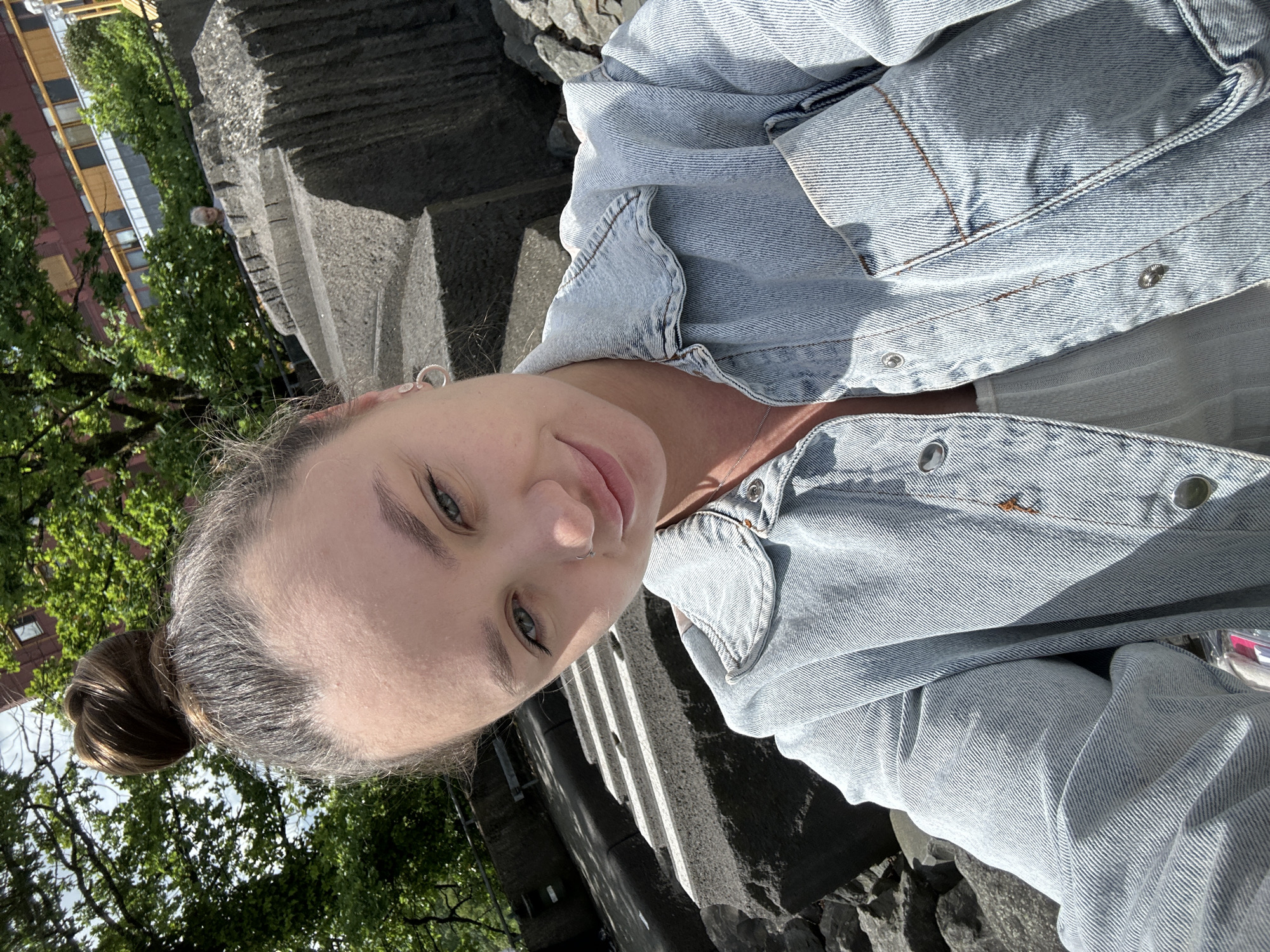 This is a close-up, selfie-style photograph of a young adult female taken outdoors, in what is identified as Gummersbach, Germany.
The subject has dark brown hair styled into a high bun atop her head, with some lighter, possibly graying, strands visible at the hairline. Her eyes are light-colored, and she gazes directly at the camera with a slight, closed-mouth smile. A small silver stud is present in her left nostril. She wears a light-wash denim jacket over a lighter-colored, possibly white or light pink, top. The denim jacket features silver-colored snap buttons, visible stitching, and a breast pocket on the wearer's right side. A small, orange-brown accessory or charm hangs from one of the jacket's front buttons. A thin silver-colored necklace is visible around her neck, and a small silver hoop earring is visible on her left ear.
The background consists of large, irregular grey stone blocks or steps on both the left and right sides, exhibiting rough textures and horizontal grooves. These structures appear to be part of a landscaped public area. Dense green foliage from trees is visible in the upper left and center background. On the far right, a multi-story building with reddish-brown walls and rectangular windows, along with yellow horizontal architectural elements, can be seen. Partially visible over a distant barrier or fence in the upper right background is an individual with gray hair, observing the scene. A dark railing runs along the top of one of the stone structures on the left. The scene is lit by natural sunlight, casting defined shadows, particularly on the denim jacket and stone elements. Rocky ground is visible in the immediate lower foreground.