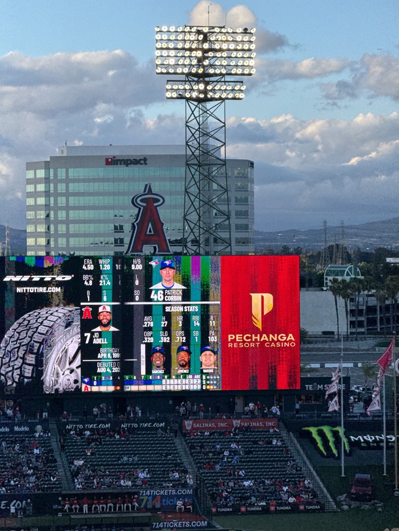 The image captures a vibrant scene at a baseball stadium, likely Angel Stadium in Anaheim, United States, under a cloudy, late afternoon or early evening sky. The focal point is a massive video scoreboard displaying game information and advertisements, backed by a large building with the "impact" logo and a prominent red Angels logo. A tall stadium light tower stands in front of the building, illuminating the field.
The scoreboard features player statistics, showing "46 PATRICK CORBIN" (a pitcher, possibly for the Toronto Blue Jays given the logo) with ERA, WHIP, H/9, BB%, and K% stats. Below, "JO ADELL" (an Angels player, #7) is highlighted with his birthday, debut date, and season stats including AVG, H, HR, RBI, R, OBP, SLG, OPS, SB, and PA. Images of other players like "SANCHEZ," "JIMENEZ," and "VARSHO" are also visible. On the right side of the scoreboard is a large red section with the "PECHANGA RESORT CASINO" logo.
Below the scoreboard, sections of stadium seating are visible, filled with numerous spectators. Various advertisements and logos are plastered throughout the stadium, including "NITTO TIRE.COM," "SALINAS, INC.," "714TICKETS.COM," "Modelo," "TUNDRA," and a large "MONSTER" (Monster Energy) logo. The overall setting is that of an active professional baseball game, with bright lights and digital displays creating an energetic atmosphere despite the cloudy conditions.