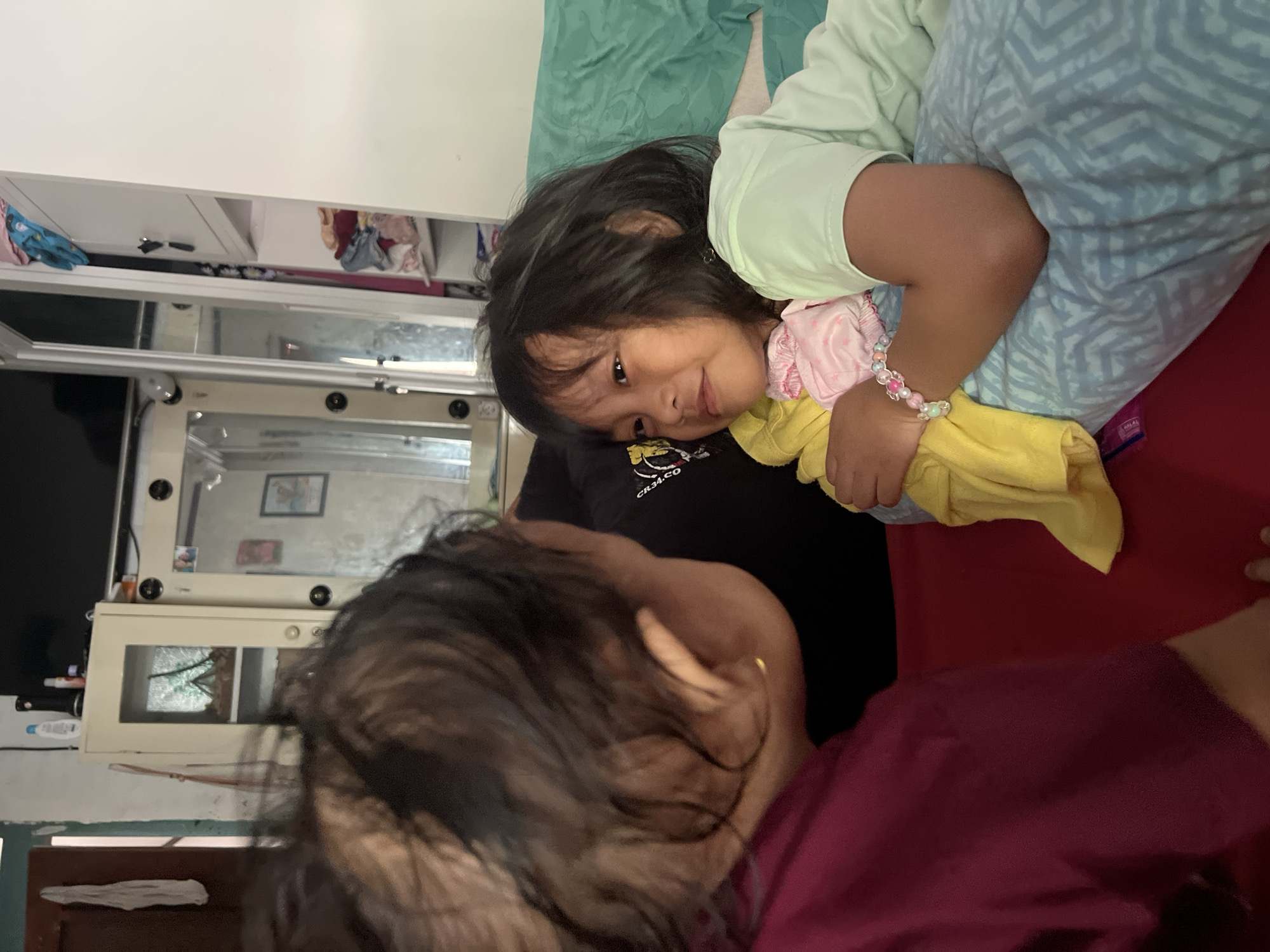 In Kecamatan Pati, Indonesia, an indoor scene captures two young girls interacting. One girl, wearing a maroon shirt, is partially visible from behind, her head turned towards the other. The second girl, with dark hair and wearing a light green shirt, is lying on a bed with blue patterned bedding and a teal blanket. She is holding a yellow cloth and a pink item. She has a bracelet on her left wrist. In the background, a large mirrored cabinet is visible, reflecting parts of the room. The environment appears to be a bedroom or living area, with household items and clothing visible on shelves.