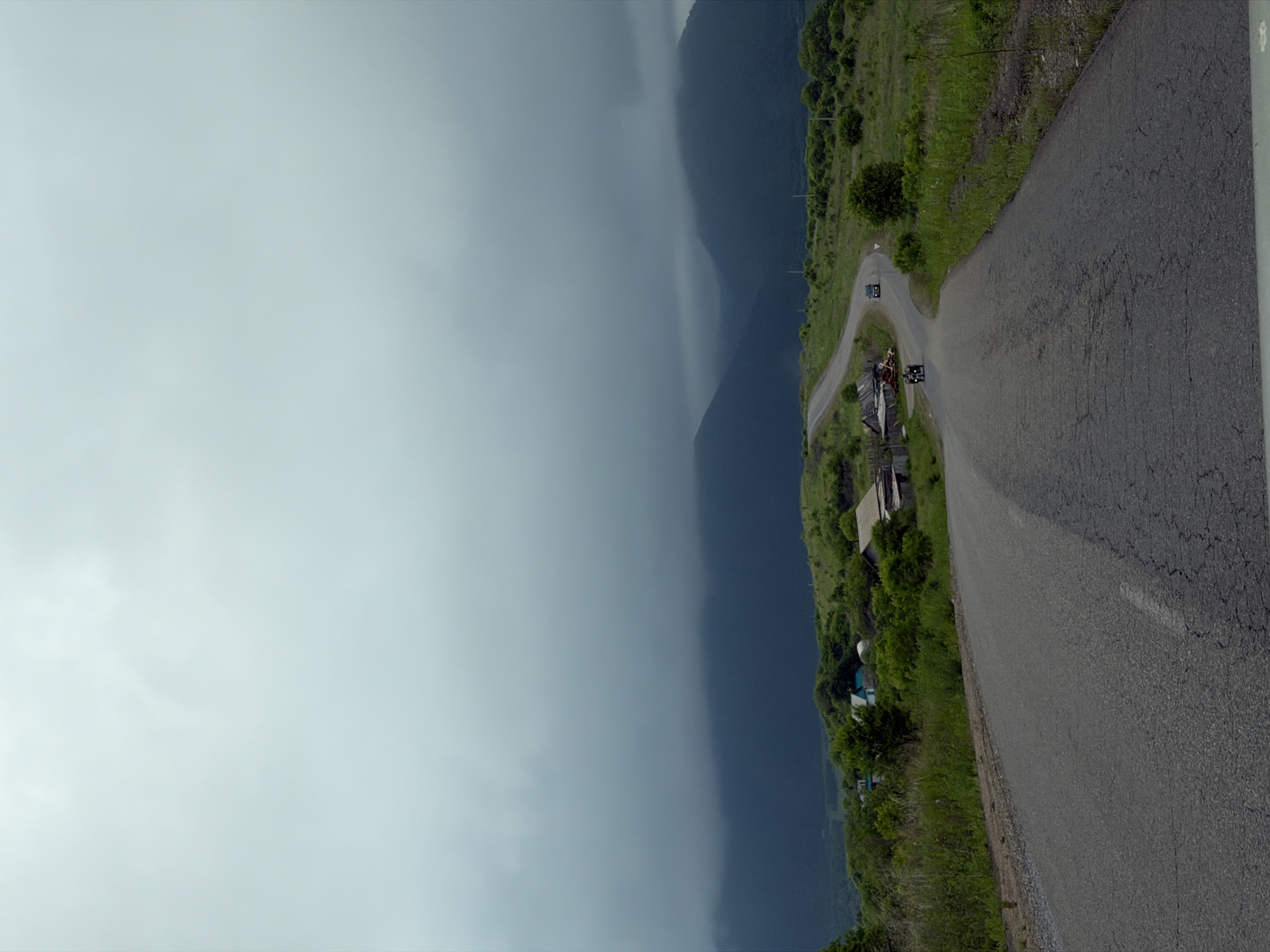 The image depicts a winding asphalt road leading through a rural landscape with rolling green hills and distant, dark mountains shrouded in heavy, overcast skies. On the left side of the road, several small, rustic buildings and structures are visible, suggesting a small settlement or homestead. Further along the road, two vehicles are present: a dark sedan in the distance and a three-wheeled motorcycle or ATV closer to the foreground, with two individuals on it. The vegetation consists of grasses and trees, typical of a temperate climate. The overall atmosphere is somber due to the cloudy weather. The location is identified as Теплоозёрск, Russia.