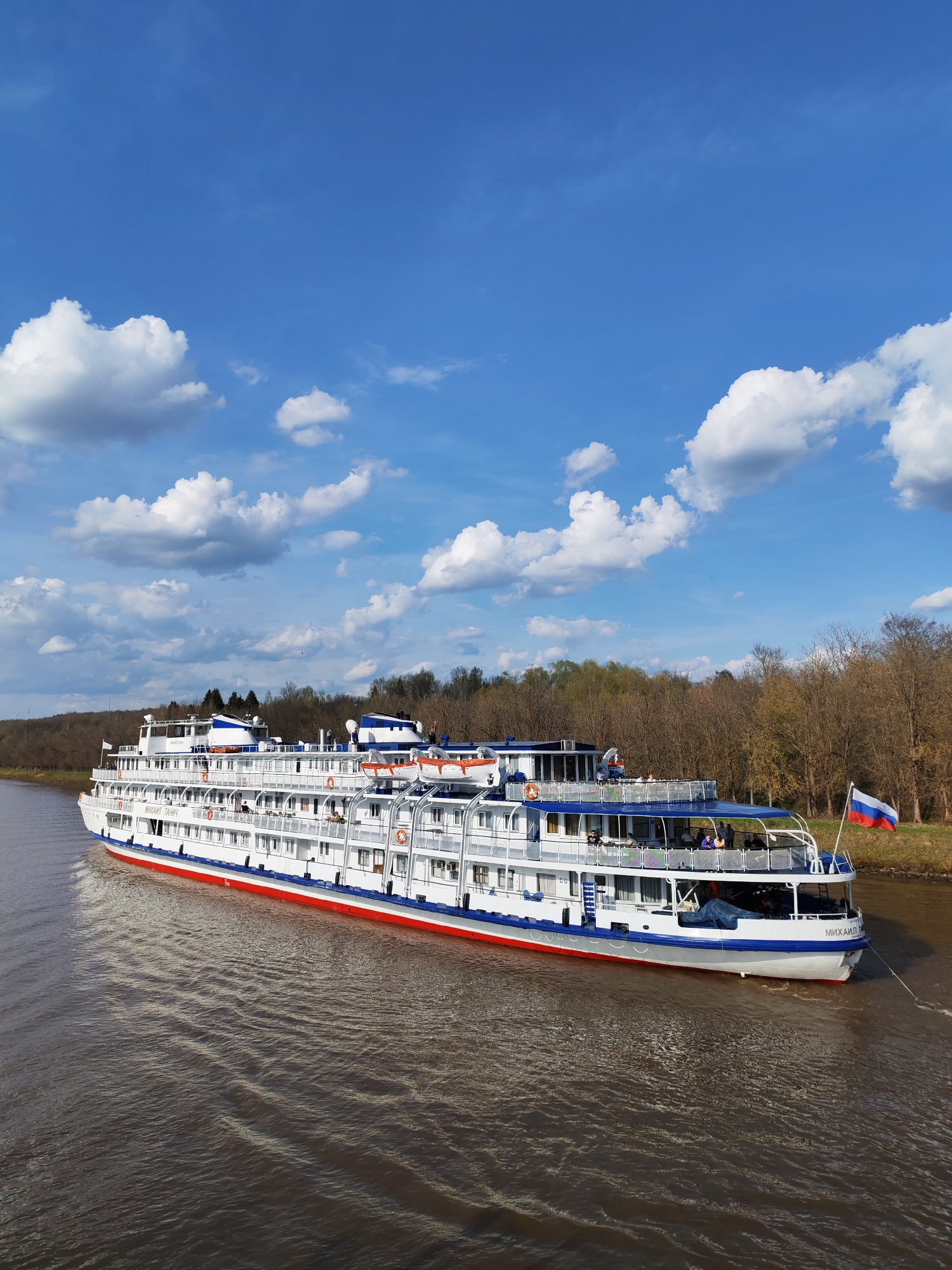 A medium-shot captures a multi-decked river cruise ship traversing a waterway. The ship is primarily white with a red stripe near the waterline and blue accents on the railings and upper decks. A Russian flag flies from the stern. Passengers can be seen on the upper decks.
The waterway appears brown, reflecting the overcast sky. Trees line the banks of the waterway. The sky is light blue with puffy white clouds scattered throughout.
The city is Unknown, Unknown.