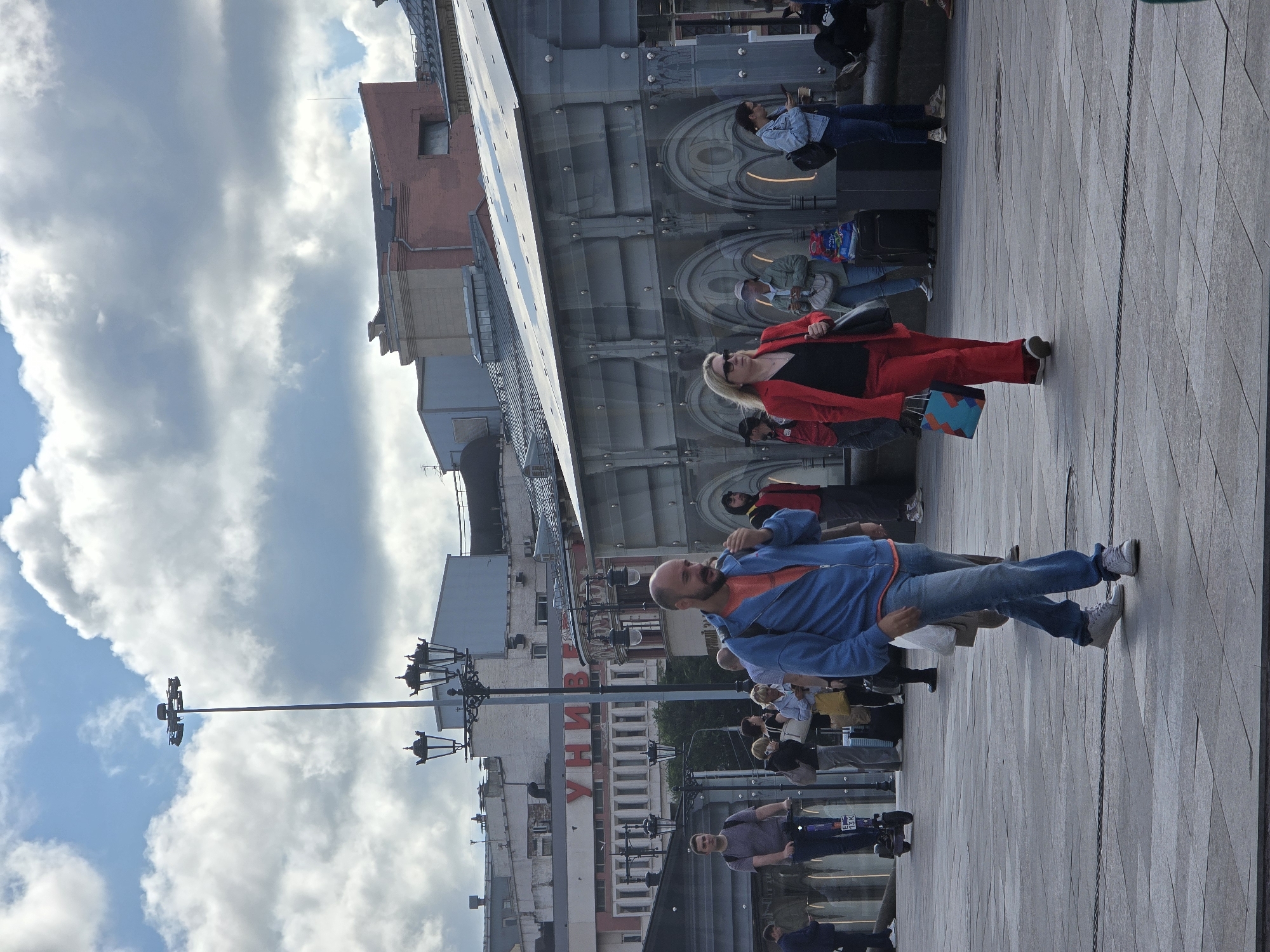 The image depicts a street scene in Moscow, Russia. In the foreground, a bald man with a beard walks towards the viewer, wearing a blue jacket over an orange t-shirt and blue jeans. He appears to be eating something. To his right, a woman in a red suit walks away from the viewer, carrying a shopping bag. Behind them, a man on an electric scooter rides towards the left side of the frame. In the background, several people are visible, some walking and others standing. Buildings with prominent signage, including the Cyrillic letters "УНИВЕР" (likely part of "Универмаг," meaning department store), are visible, suggesting a commercial area. Streetlights and a tall lighting mast are also present. The sky is partly cloudy. The scene appears to be taking place in an open public space, possibly a pedestrianized area or a plaza.
