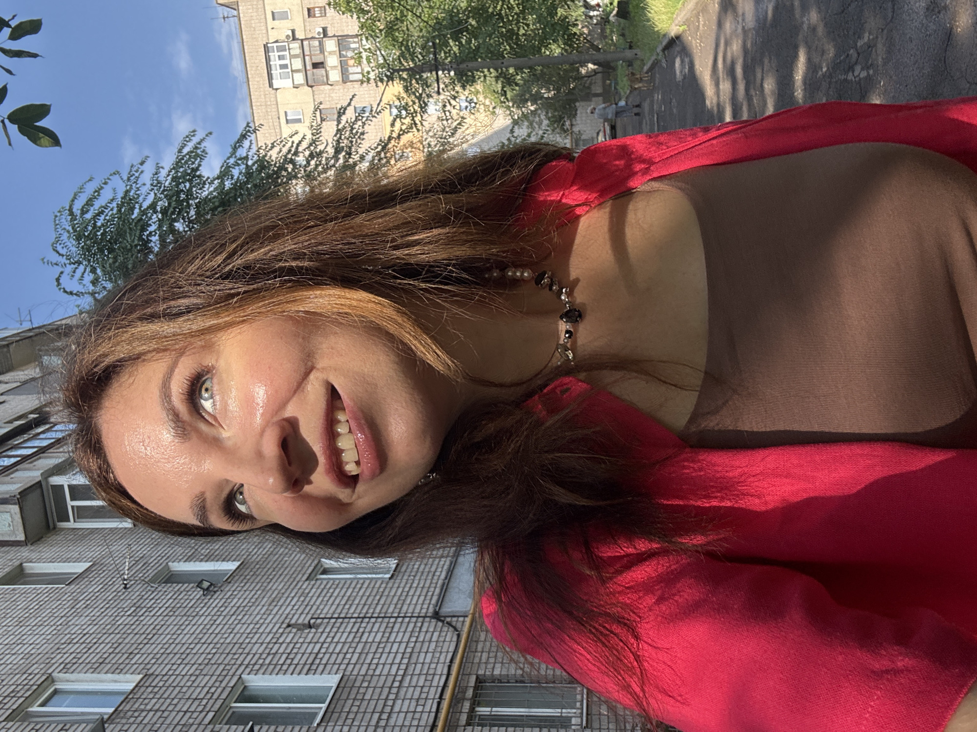A young woman with brown hair and blue eyes is shown from the chest up, looking towards the sky with a smile. She is wearing a pink cardigan over a brown tank top and a necklace. The background consists of an apartment building on the left and trees and another apartment building on the right, with a clear blue sky above. The scene appears to be an outdoor urban environment.