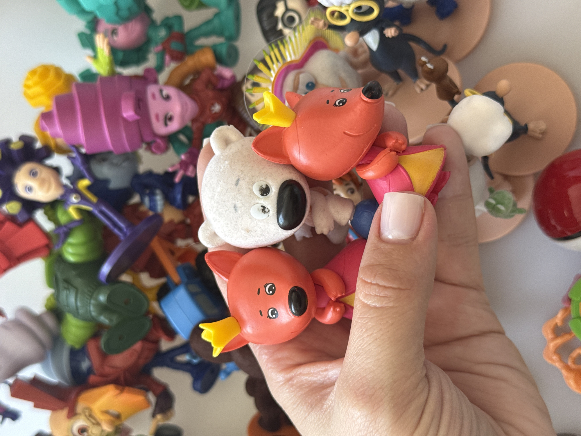 This is a close-up, eye-level, indoor photograph of a hand holding three small, plastic toy figures. The figures are stylized, cartoon-like characters with simplified features. The figure in the center is a white, fluffy bear with black eyes and nose. To its left is a red fox-like creature wearing a yellow crown and a pink dress with a yellow trim. To the right of the bear is another red fox-like creature, also wearing a yellow crown and a pink dress with a yellow trim. The hand holding the figures is a fair-skinned adult hand with a pale pink fingernail polish on the visible fingernail. The background is filled with a multitude of other similar toy figures, mostly in muted colors, suggesting a collection of toys. The lighting is bright and diffused, indicating an indoor setting. There is no information in the image to suggest a location in Lyubertsy, Russia.