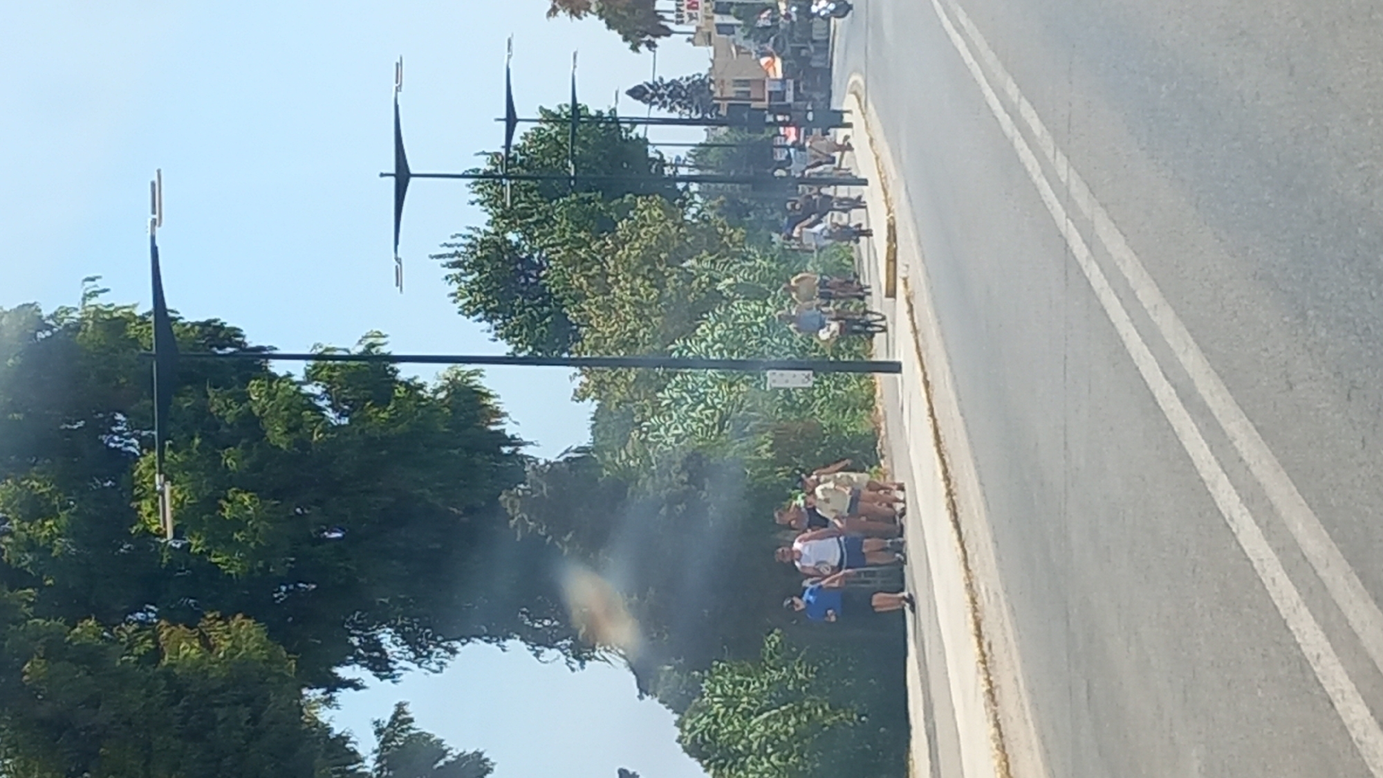 The image shows a street scene in Heraklion, Greece, on a sunny day. A group of people are walking on the sidewalk on the left side of the road. Some are wearing casual summer clothing, and one person is wearing a blue shirt and dark shorts. Further down the sidewalk, several people are riding bicycles. On the right side of the road, there are buildings, including a sign that appears to say "SUPER MARKET". The street has a paved surface with double white lines indicating traffic lanes. Several modern streetlights with horizontal lighting fixtures are visible along the sidewalk. Lush green trees and bushes line the sidewalk, providing shade. The sky is clear and blue.