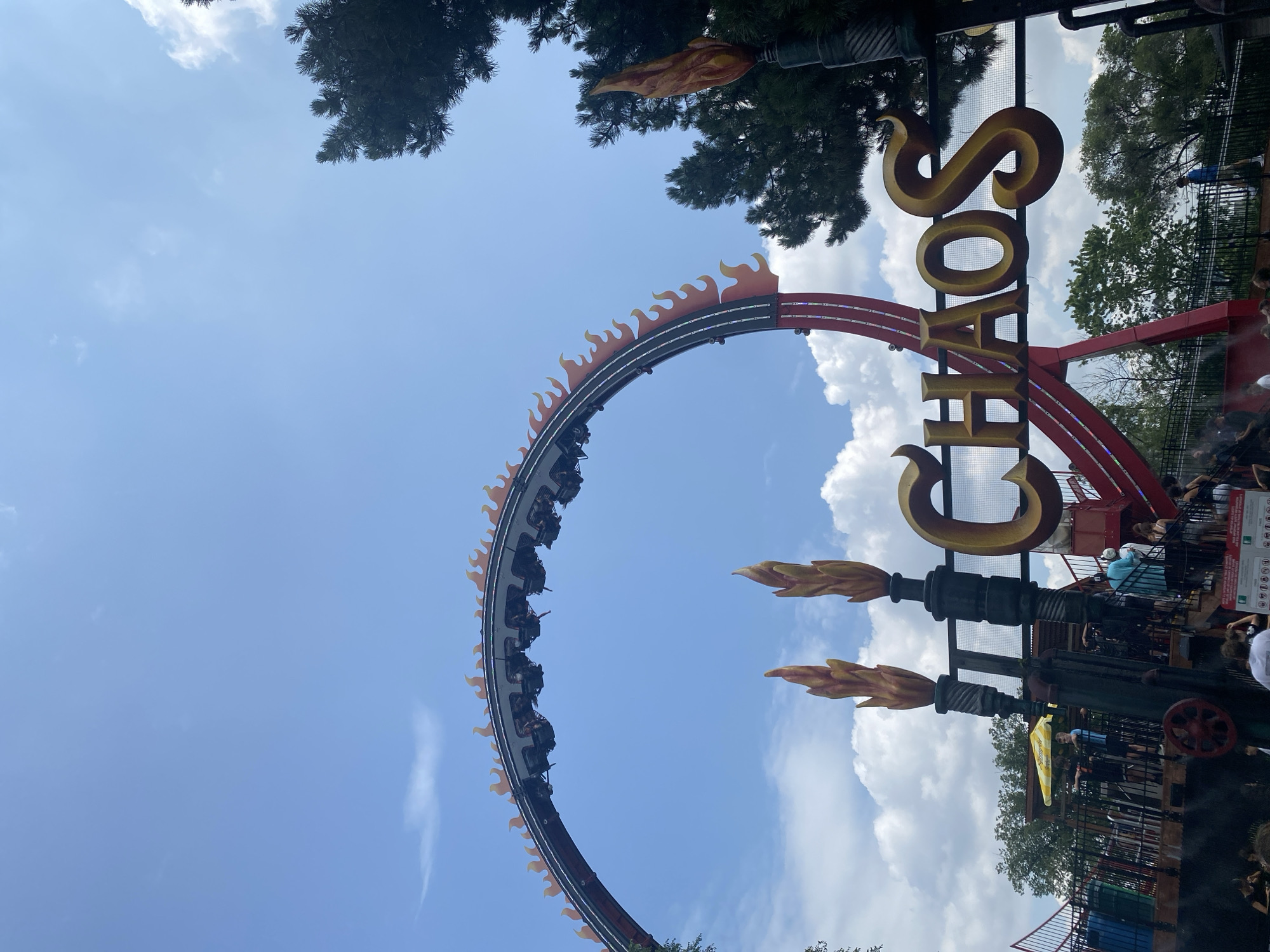 This image shows the "Chaos" roller coaster ride at La Ronde, an amusement park located on Île Sainte-Hélène in Montreal, Canada. The ride features a large loop with flames decorating its edges, and a train of cars carrying passengers is suspended upside down at the apex of the loop. In the foreground, a sign with the word "CHAOS" in large, golden letters is visible, flanked by two torch-like structures with flame designs. People are gathered on the ride's platform and in the queue area, with some standing on stairs. The environment is sunny with a bright blue sky and scattered clouds, and trees are present in the background.