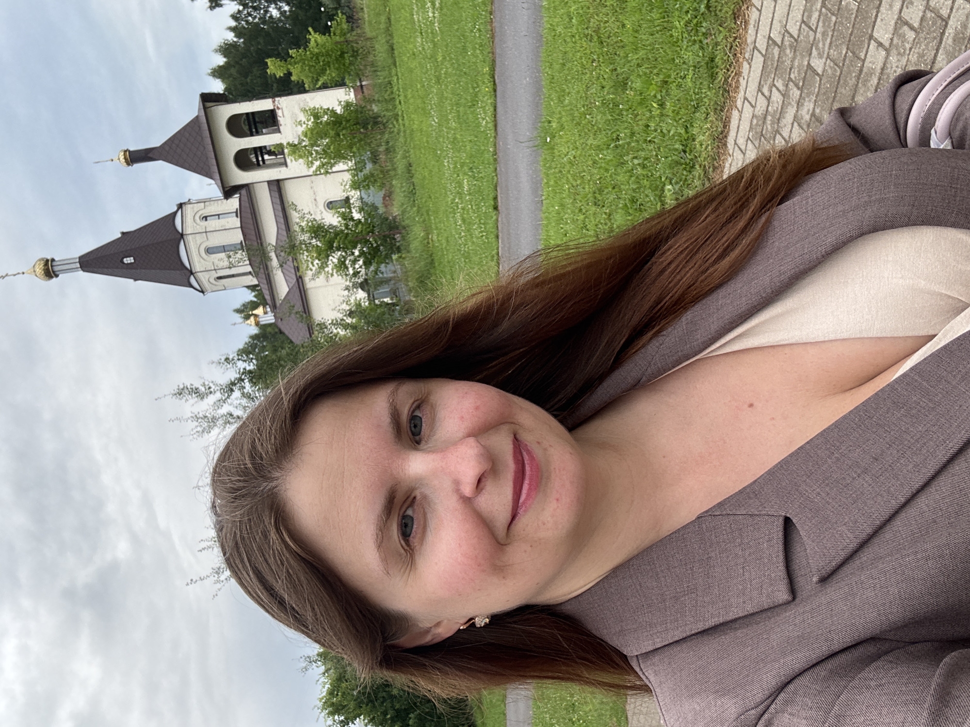 A woman with long, brown hair, wearing a taupe blazer over a light-colored V-neck top, smiles at the camera. Her left earlobe sports a small, dangling earring. She is standing outdoors with a paved walkway in the foreground. Behind her, a light-colored church with a dark-brown roof and a bell tower is visible. The church has a golden dome. Green grass and trees surround the building, and the sky is overcast. The location is the Church of the Intercession of the Most Holy Theotokos in Konakovo, Russia.
