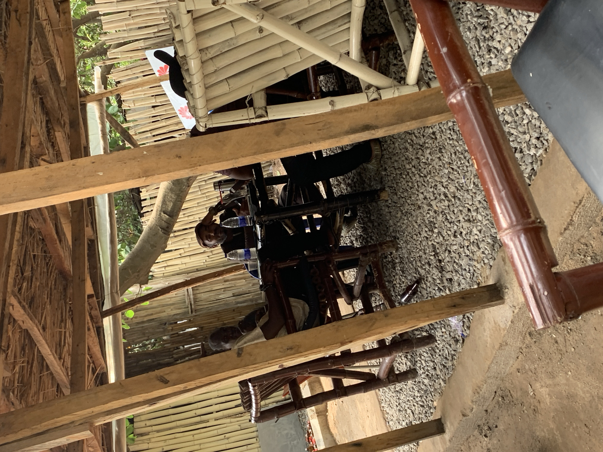 The image depicts a group of people seated on chairs under a rustic, open-air structure constructed primarily of bamboo and wood. The roof is made of tightly packed bamboo poles, and the walls, where visible, appear to be woven bamboo or straw. Sunlight filters through the gaps, suggesting it is daytime. The ground is covered with small grey pebbles.
There are at least three individuals visible, appearing to be men, dressed casually. One person is leaning back in a chair, looking towards the camera. Another person is seated and appears to be holding a bottle or can. A third person is partially visible, also seated. They are positioned around tables or a central structure, possibly a bar or dining area. Various bottles, including some with water and possibly beer, are scattered around. The overall impression is a relaxed, informal gathering, possibly at a casual eatery or a shaded outdoor seating area in Abuja, Nigeria. The lighting suggests it could be a sunny day, but the specific time is difficult to determine. The mood seems casual and unhurried. No discernible text is visible in the image.