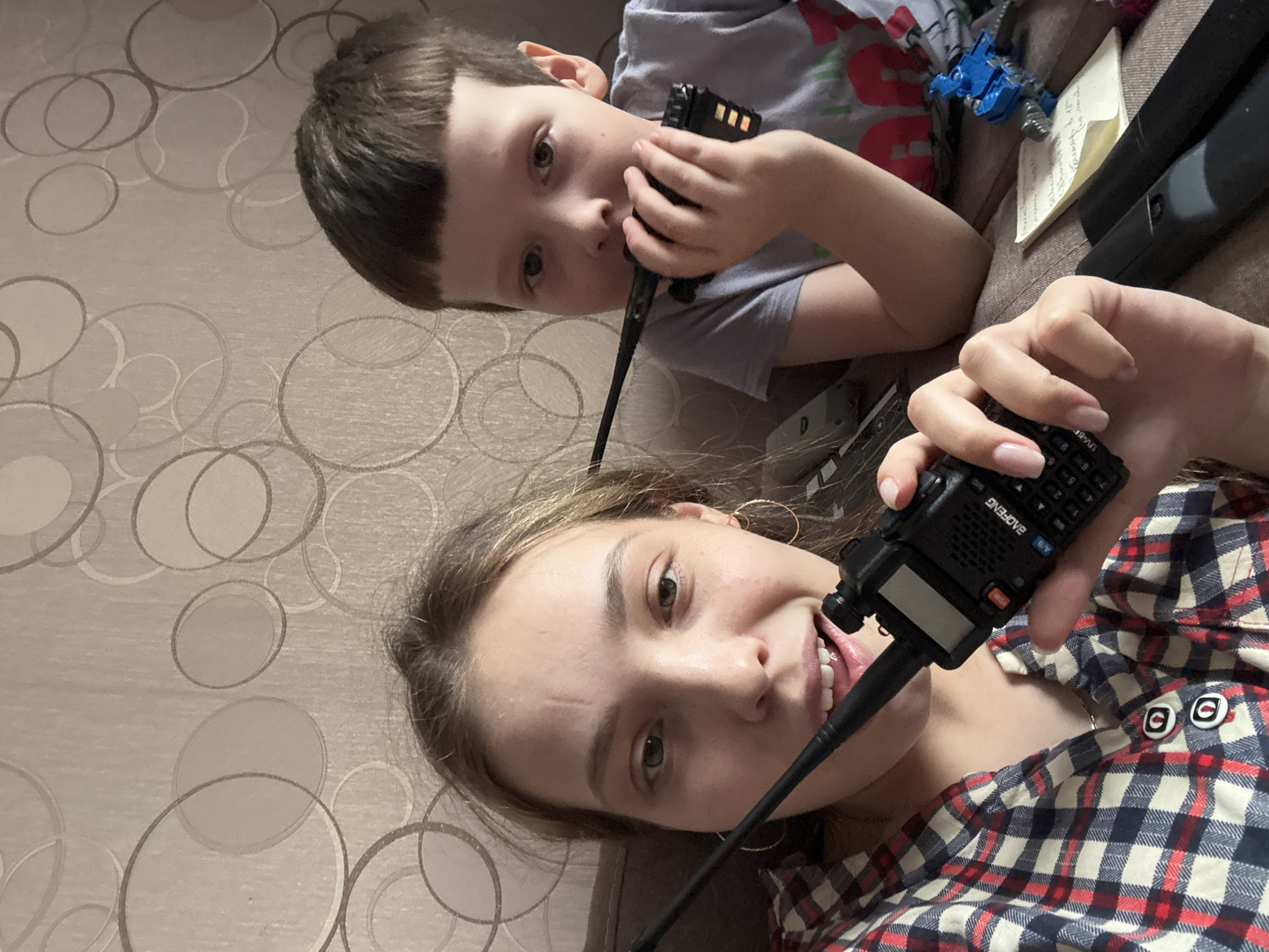 Two individuals, a child and an adult, are pictured holding two-way radios. The child is on top, wearing a grey shirt, and holding the radio to his mouth. The adult is below, wearing a red and blue plaid shirt, also holding a radio to their mouth. The background shows a wall with a circular pattern. The location is Кемуль, Russia. Other objects are visible in the background, including a blue toy and a notepad.