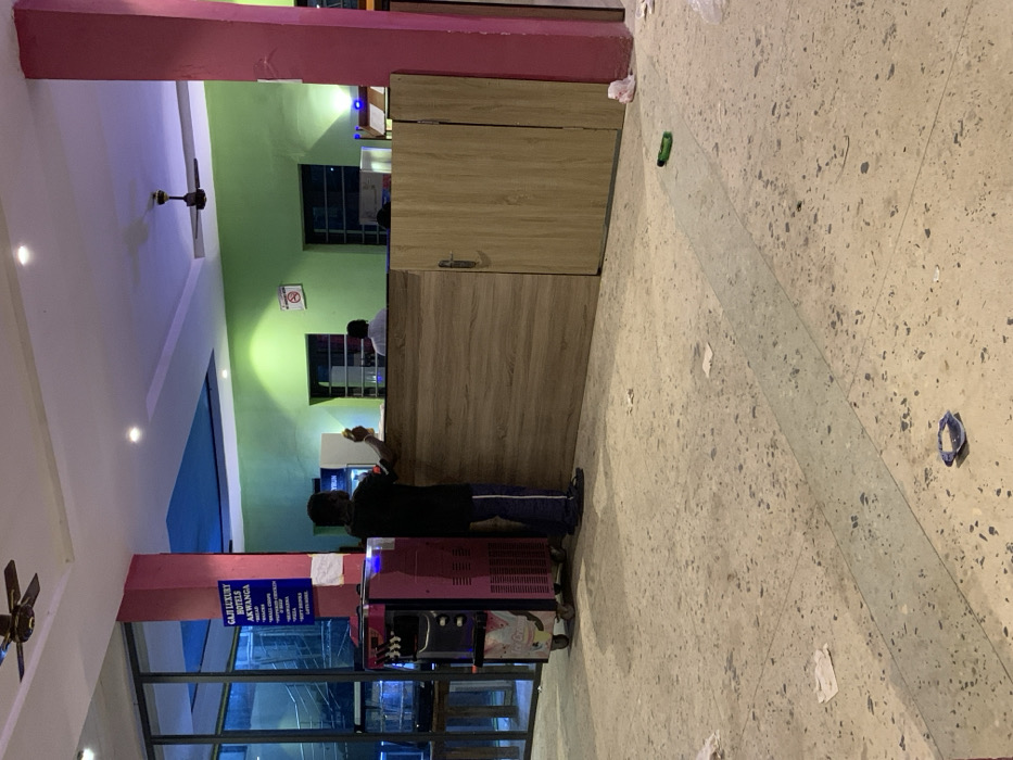 The image depicts the interior of Gaji Luxury Hotels, located in Akwanga, Nigeria. The scene is centered on a service or reception counter with a wooden-patterned laminate finish. A male individual, viewed from behind, stands on the customer side of the counter, wearing a dark t-shirt and dark trousers with white side stripes, holding a white, rectangular object in his right hand. To his left, a pink, wheeled ice cream or frozen yogurt machine with multiple dispensing spouts is positioned. Attached to a prominent pink structural column to the left of the counter is a blue sign displaying menu items including "GAJI LUXURY HOTELS AKWANGA", "BREAD", "SMALL CHOPS", "CHICKEN & CHIPS", "SHAWARMA", "COFFEE", and "SOFT DRINKS". Behind the counter, a second male individual, wearing a white shirt, is partially visible. The interior walls are painted lime green, featuring a "NO SMOKING" sign. The ceiling is multi-tiered with blue and white sections, illuminated by recessed lighting, and includes a single ceiling fan. The light-colored, speckled floor exhibits scattered debris, including several pieces of paper, a small green cylindrical object, and a blue plastic item. A large glass partition is visible on the far left.