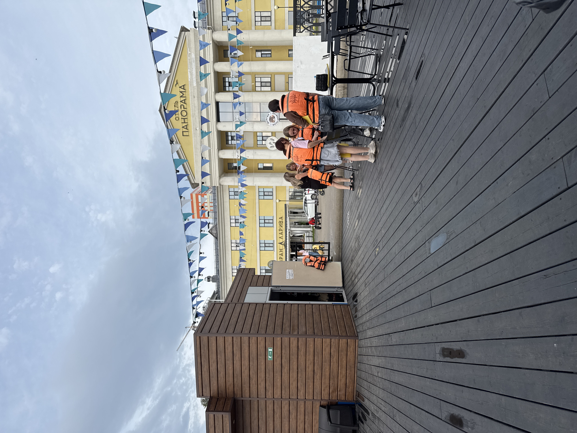 This image shows a group of people wearing orange life vests gathered on a dark wooden deck in front of a yellow building labeled "ОТЕЛЬ ПАНОРАМА" (Hotel Panorama). The building has large columns and is decorated with triangular flags. To the left, there is a wooden structure with an open doorway. People are interacting with each other and appear to be preparing for an activity. The environment suggests an outdoor area, possibly a waterfront or recreational space, in Izhevsk, Russia.