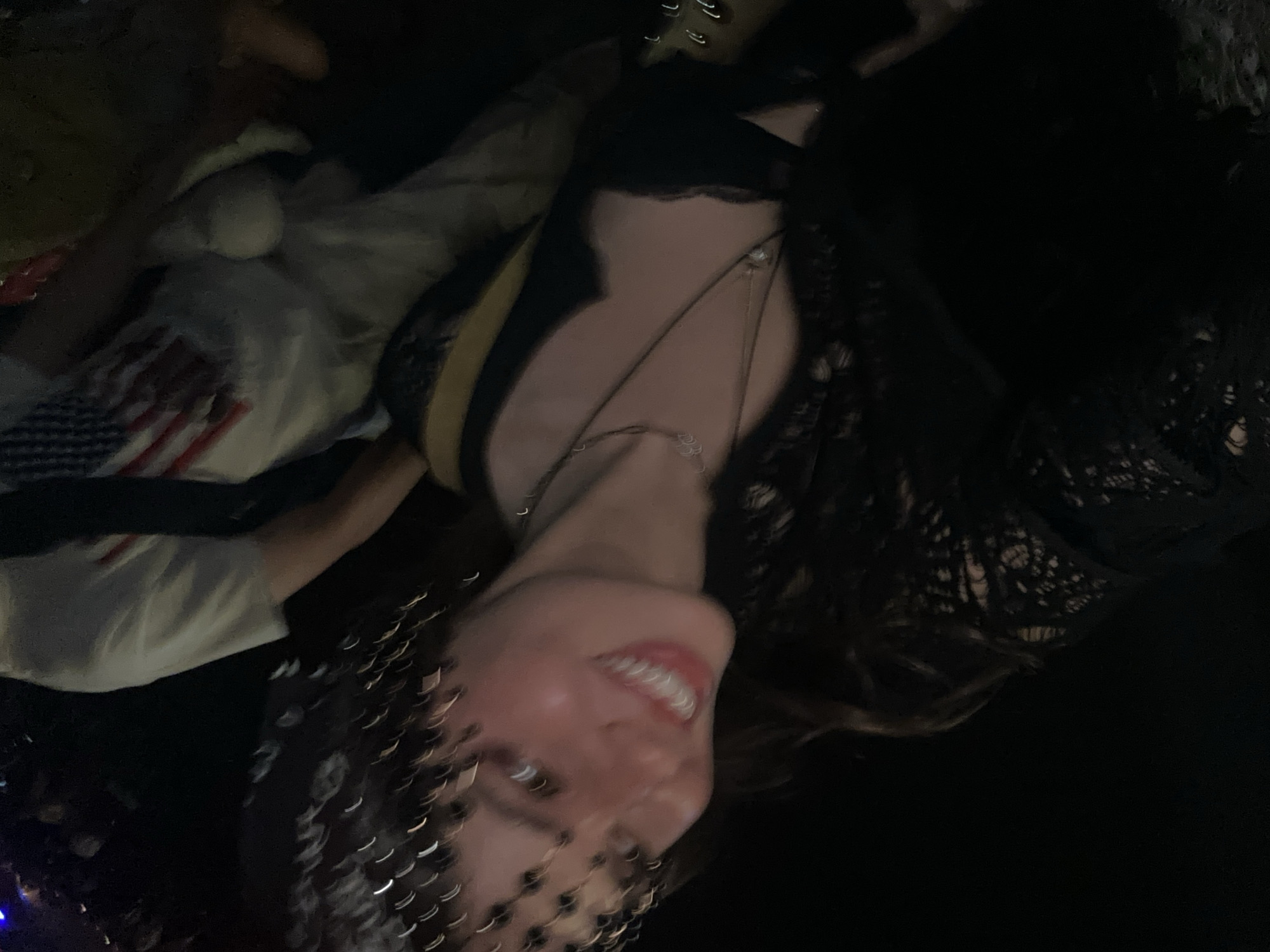 A low-angle, slightly blurry image shows a woman with light skin, wearing a black lace top and multiple necklaces, smiling at the camera. She has a headpiece that appears to be made of small, reflective beads or sequins. To her left, someone is wearing a white t-shirt with the image of the American flag on it. The environment appears to be dark, suggesting the image was taken at night. Given the prompt's mention of Indio, United States of America, and the clothing and accessories visible, it is plausible the image was taken at a music festival, possibly the Coachella Valley Music and Arts Festival, held annually in Indio.