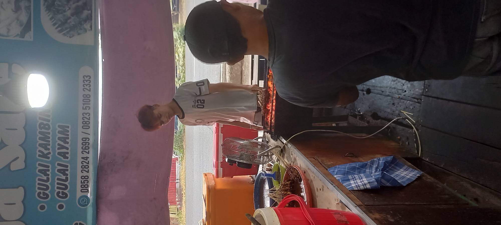 In Melayu, Indonesia, an outdoor food stall is captured in a medium shot. The stall has a purple awning overhead, and a blue sign with white lettering is visible, listing menu items like "GULAI KAMBING" and "GULAI AYAM," along with phone numbers: "0858 2824 2699 / 0823 5108 2333."
A young man with reddish-brown hair, wearing a white t-shirt with "02" printed on it, is tending to a grill. He is looking down at the food on the grill, which appears to be satay or skewers cooking over hot coals. A fan is positioned on the counter to his left. In front of him, there are two large coolers, one orange and one red, and a blue bowl containing satay skewers and a brush. To the right of the frame, the back of another person wearing a dark t-shirt and a black cap is visible, facing the grill. The counter is made of wood, and the floor beneath is also wooden planks. The scene suggests a busy food preparation area.