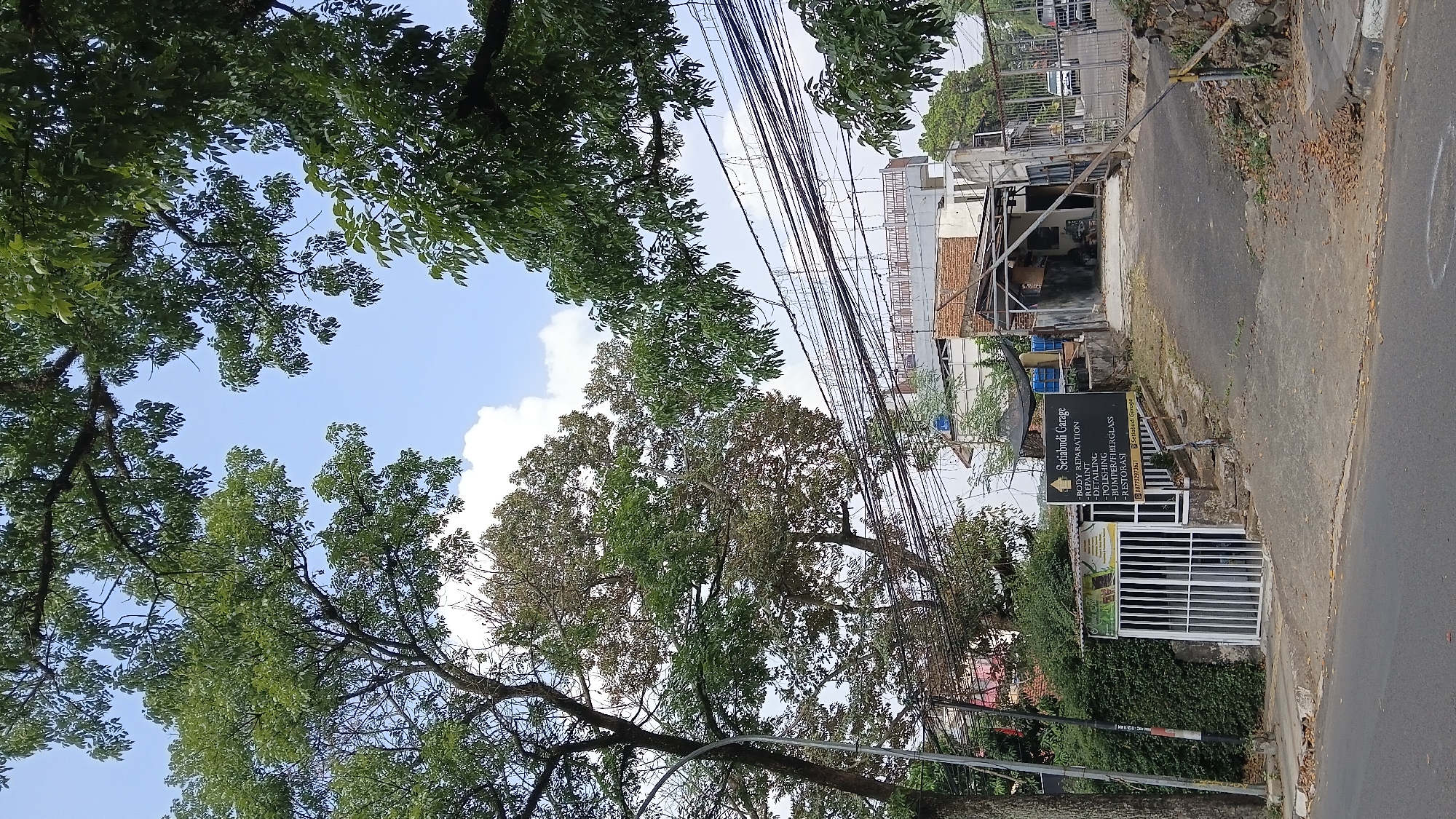 A low-angle exterior shot captures a scene in front of a garage in Bandung City, Indonesia. Large trees with green leaves dominate the upper portion of the frame, their branches extending across the sky, which is partly cloudy with patches of blue. In the mid-ground, a sign for "Setiabudi Garage" lists services like "BODY REPARATION," "REPAINT," "DETAILIMG," "POLISHING," "BUMPER/FIBERGLASS," and "RESTORASI." Below the listed services is a phone number and the garage's name again. The garage entrance is visible, appearing somewhat dilapidated with exposed brick and corrugated metal roofing. A white gate or door with vertical bars is on the left side of the scene, leading into a small building or shed. A paved driveway leads up to the garage. Various electrical wires crisscross the sky in the mid-ground. The ground in front of the garage is covered in scattered dry leaves. A street is visible in the foreground. There are no people or vehicles actively engaged in any activity in the immediate vicinity.