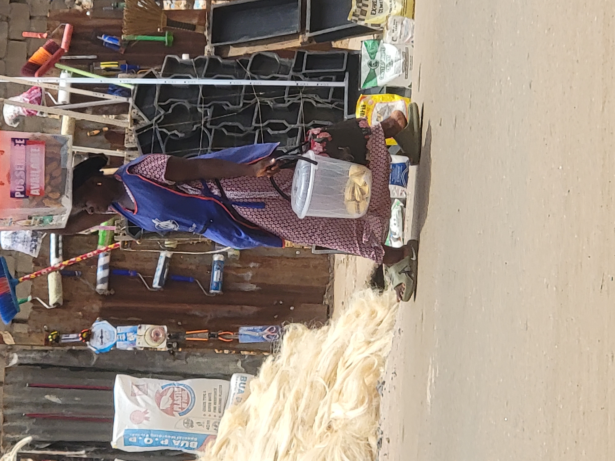 This image, rotated 90 degrees counter-clockwise, captures an outdoor scene, likely a street market or roadside stall, in Maiburiji, Nigeria, during bright daylight.
In the foreground, a person, possibly a woman, stands facing left. She wears a patterned pink and black dress or top underneath a blue vest. She has a dark head covering and light green sandals on her feet. She holds a clear plastic container filled with light-colored snacks, possibly plantain chips, suggesting she might be a vendor.
The background is rich with various goods for sale, indicating a commercial setting. These include brooms, paint rollers, and brushes hanging on what appears to be a corrugated metal or wooden wall. Other tools like pliers and scissors are also visible. On the ground, next to the person, is a large pile of light-colored, fibrous material, resembling natural fibers or yarn. Behind the person and stacked against the wall are numerous bags of products, some white, green, yellow, and brown.
Visible text on the bags includes "BUA P.O.P" and "BUA D PLASTHE" on white bags, and "CLMET", "EX-RE", "CINSTAHITE", and "SOFIA" on others. A pink sign or box in the upper left corner prominently displays "POS SEREIBE AVAILABLE." The ground is an unpaved or concrete surface. The overall impression is a bustling, informal marketplace.